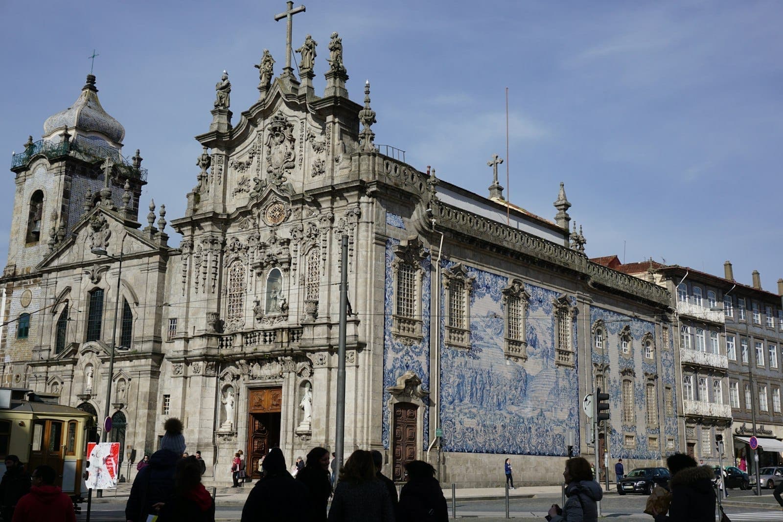 Weitwinkelperspektive auf die prachtvolle Barockfassade der Igreja do Carmo und das berühmte blau-weiße Azulejo-Kachelpaneel an der Seite, mit Menschen und Gebäuden rundherum an einem sonnigen Tag.