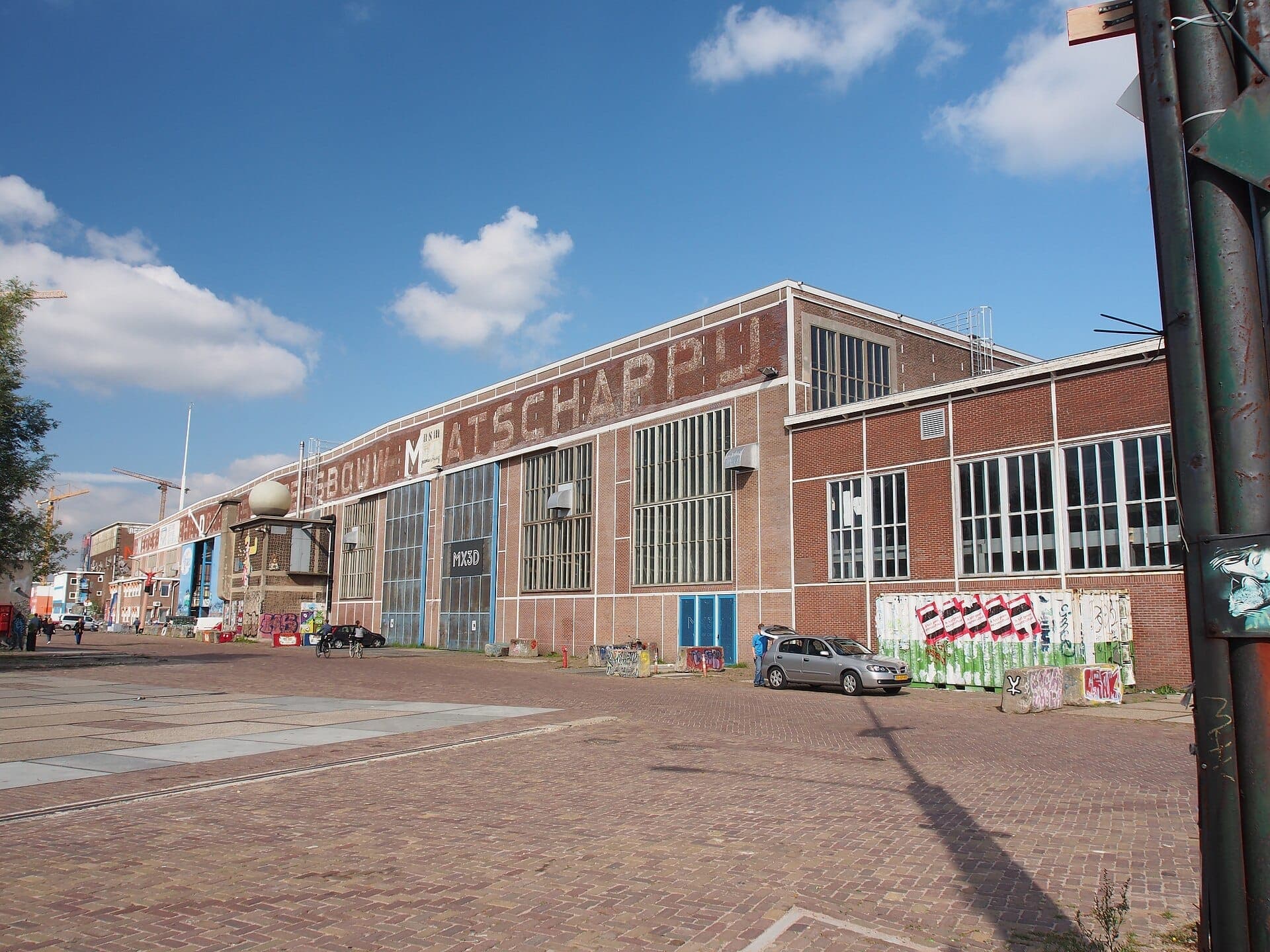 Außenansicht der Industriehalle NDSM, in der der Flohmarkt IJ-Hallen stattfindet, mit roten Backsteinwänden, großen Fenstern und vereinzelten Street-Art-Werken.