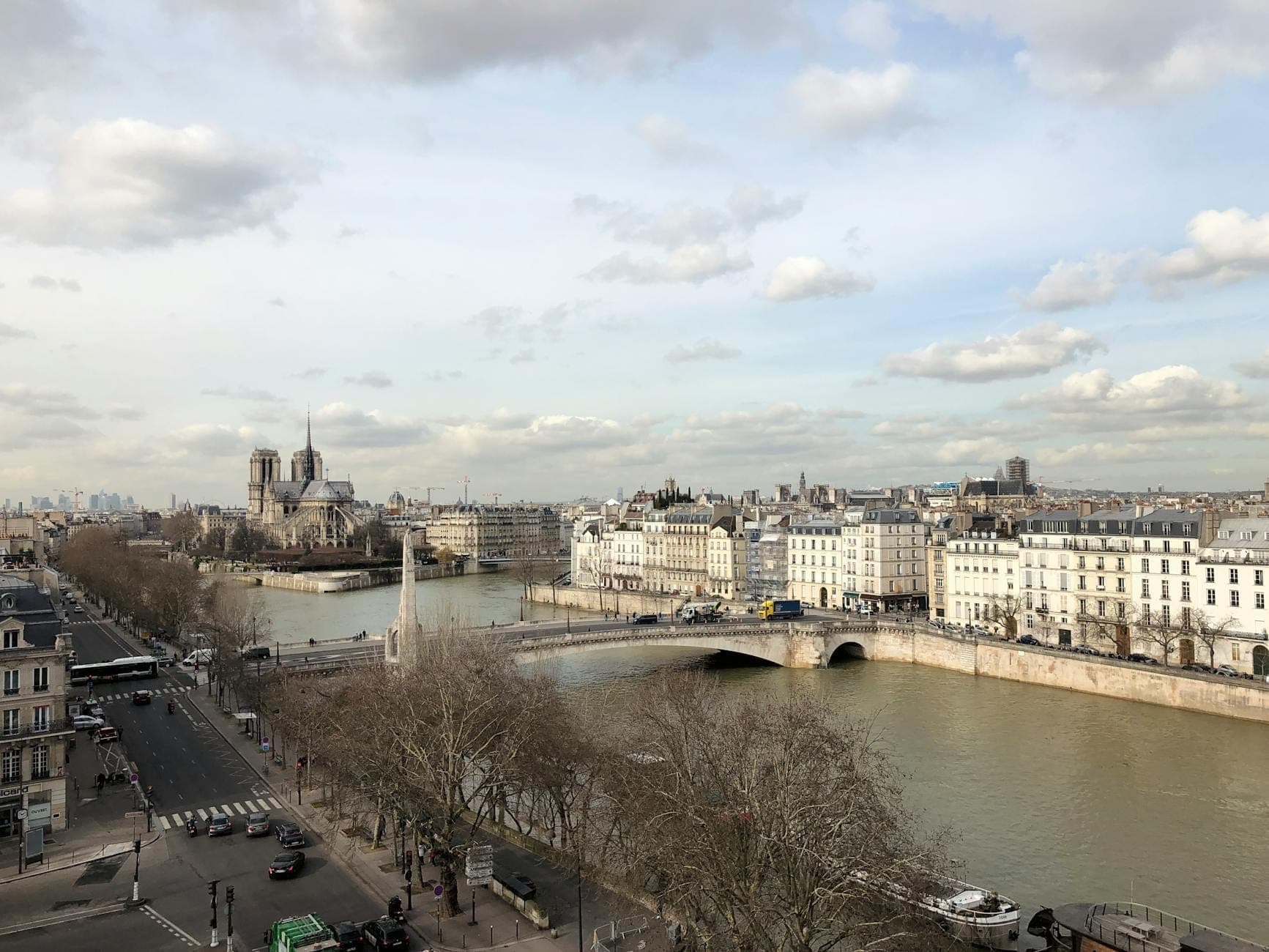 A panoramic view of Île Saint-Louis in Paris, showing elegant stone buildings, bridges, the Seine River, and Notre-Dame in the distance under a cloudy sky.