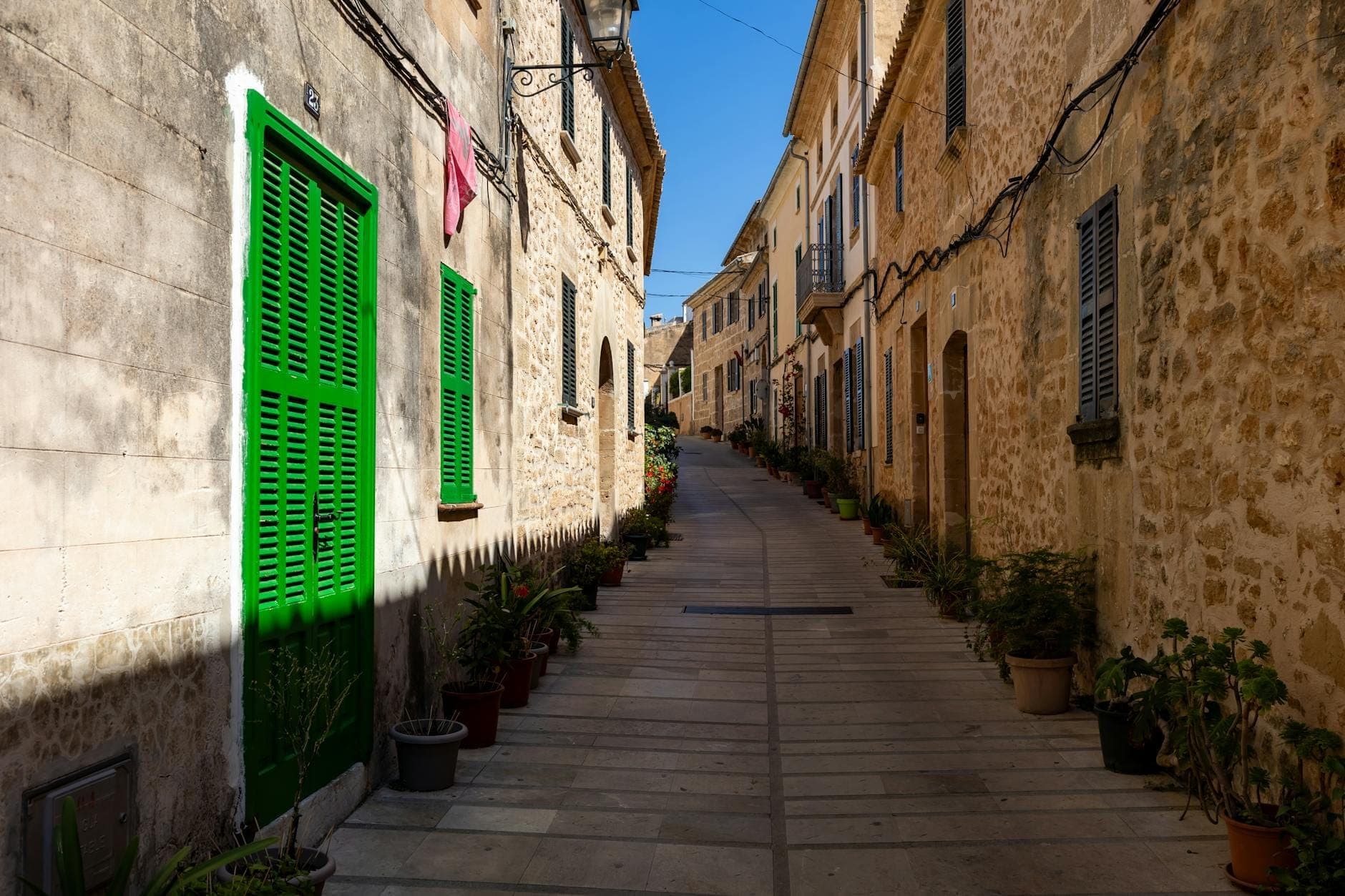 Enge Steinstraße in Es Pla, Mallorca, gesäumt von Topfpflanzen und traditionellen Häusern mit grünen und blauen Fensterläden im hellen Sonnenlicht.