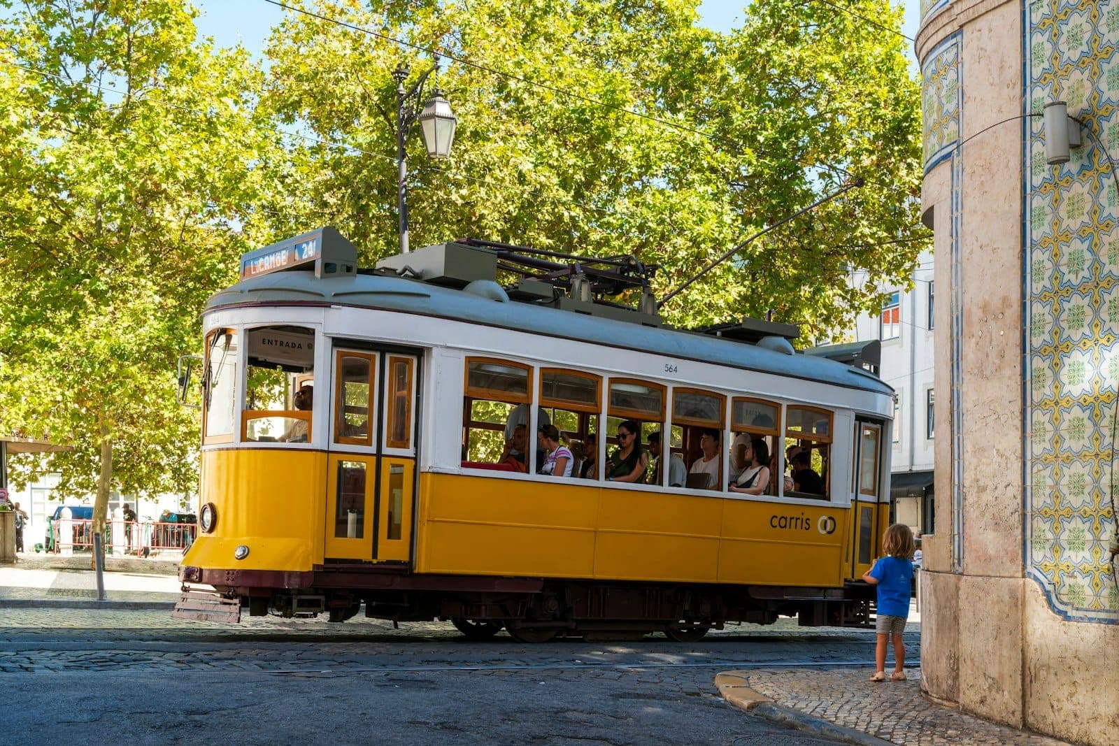 Elétrico amarelo clássico a virar numa esquina de Lisboa, com fachada de edifício azulejada e árvores frondosas ao fundo, captando a autêntica vida da cidade.