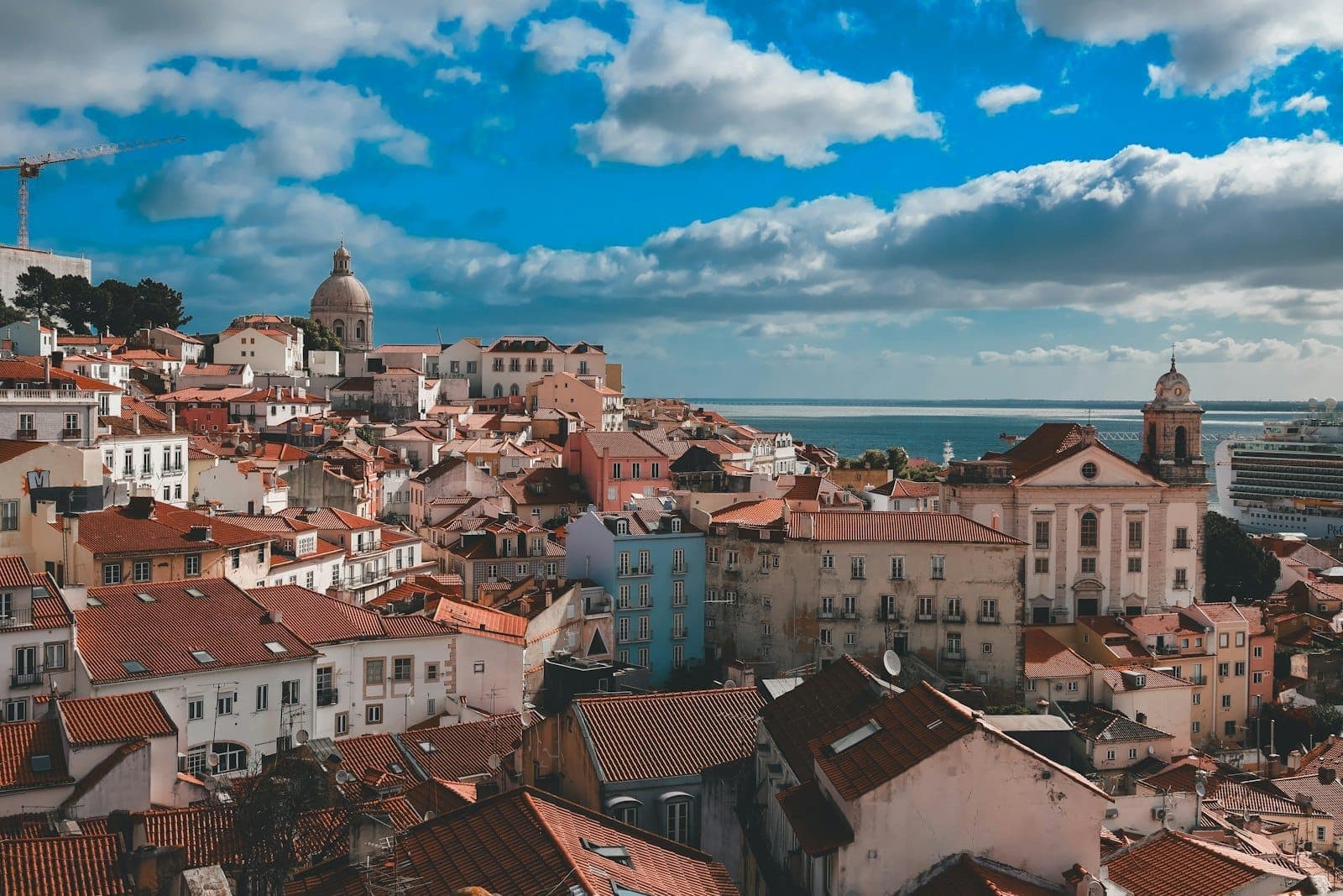 Vista panorâmica dos telhados vermelhos de Lisboa, com prédios históricos, uma igreja com cúpula e o rio Tejo sob um céu azul vibrante com nuvens esparsas.