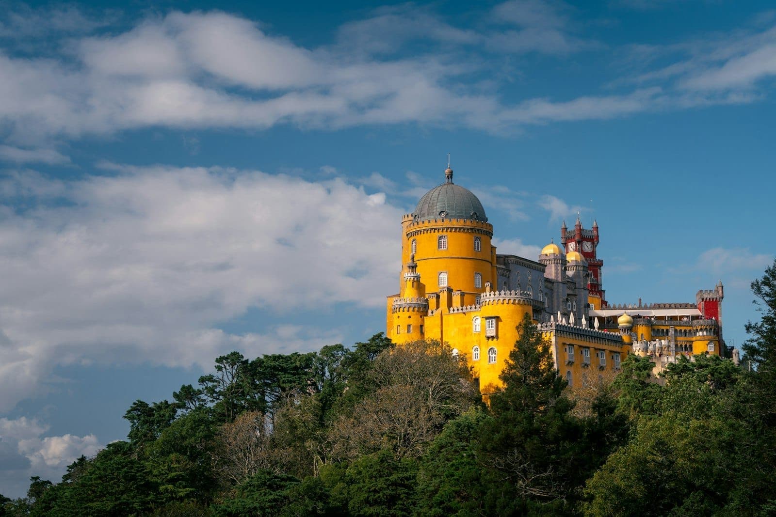 Vista ampla do colorido Palácio da Pena no topo de uma colina arborizada verde sob um céu azul com nuvens esparsas perto de Lisboa.