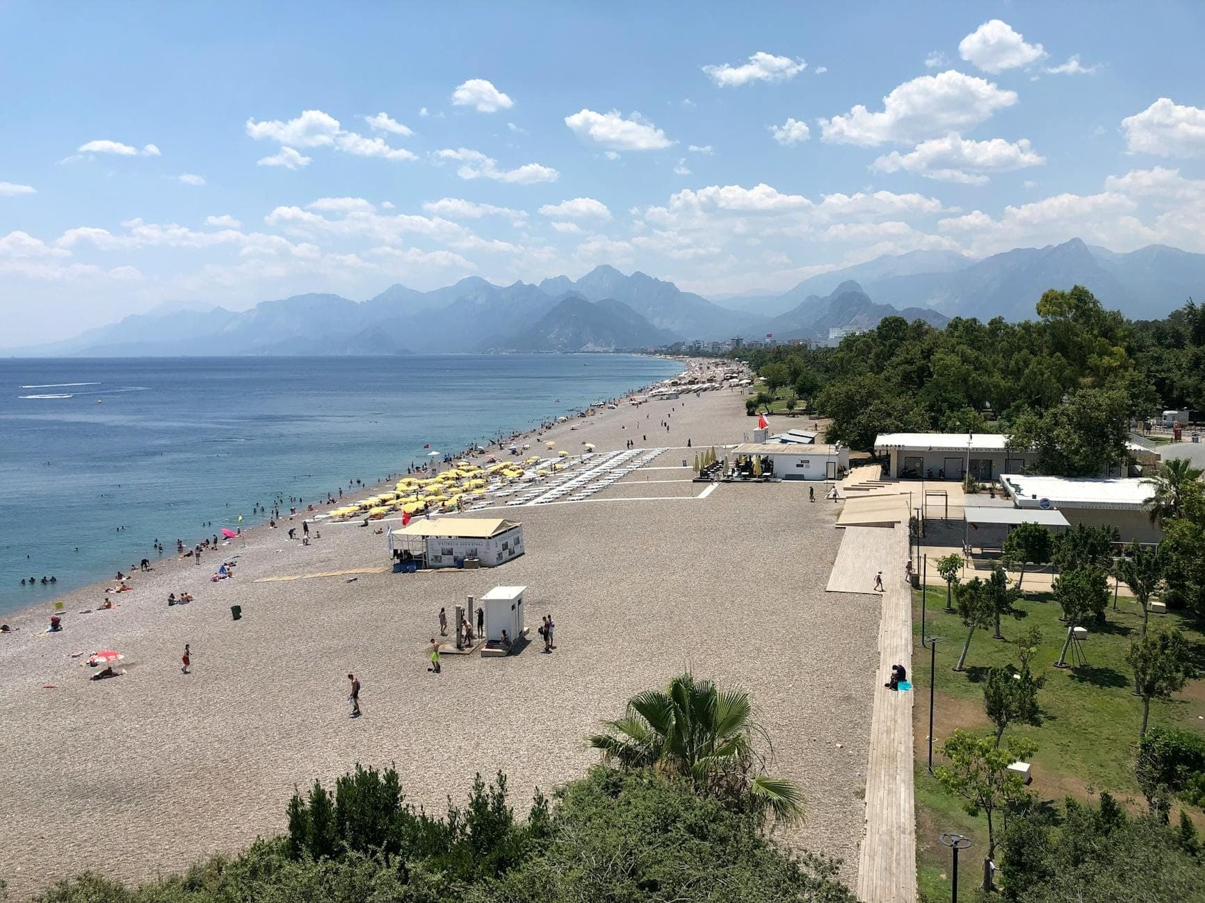 Ein Kiesstrand in Antalya mit Menschen, Liegestühlen, dem Grün eines nahegelegenen Parks und dem beeindruckenden Taurus-Gebirge im Hintergrund.