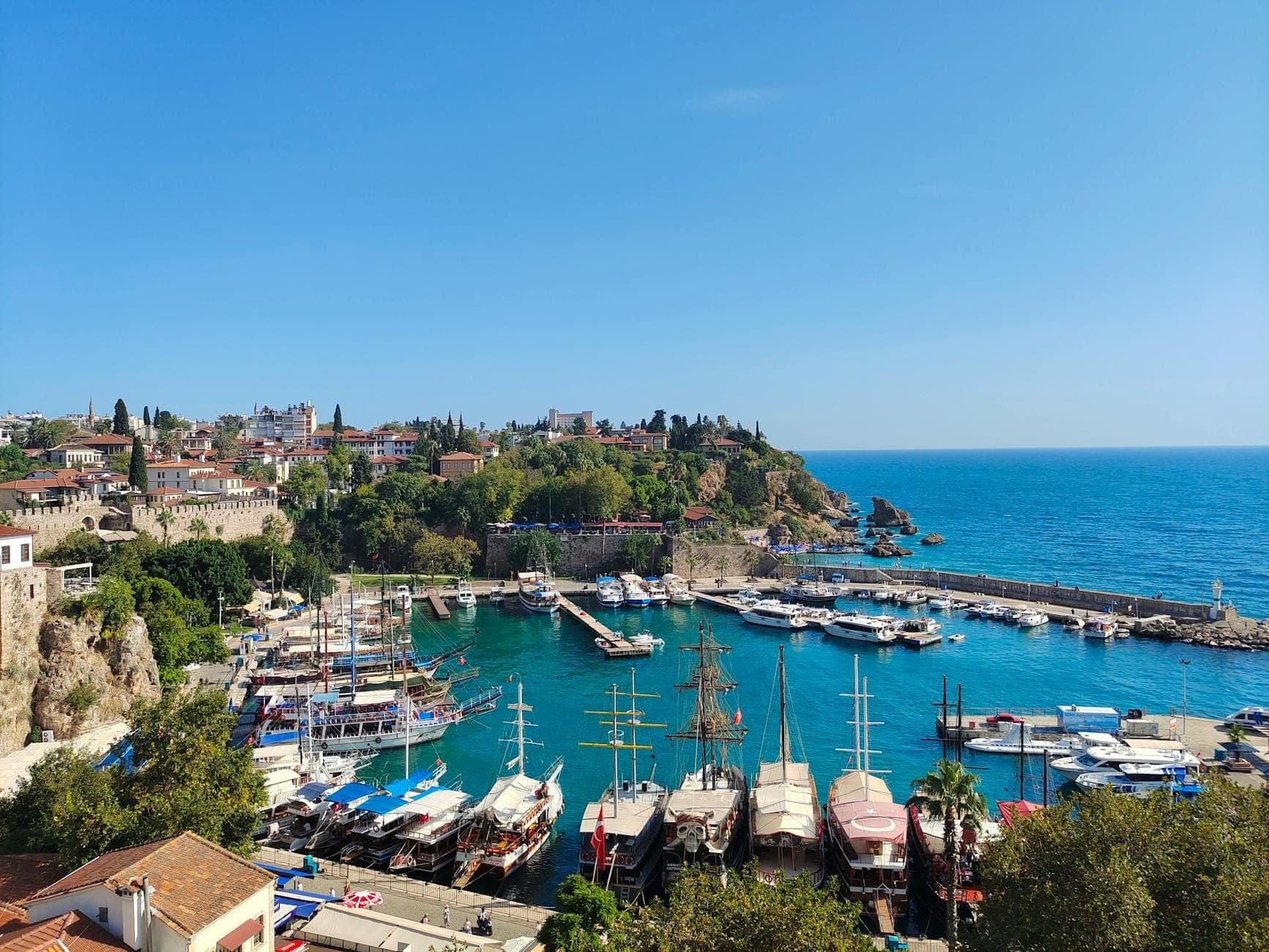 Luftaufnahme des alten Hafens von Antalya mit türkisfarbenem Wasser, traditionellen Booten, historischen Steinmauern und der Stadtsilhouette an einem strahlend sonnigen Tag.