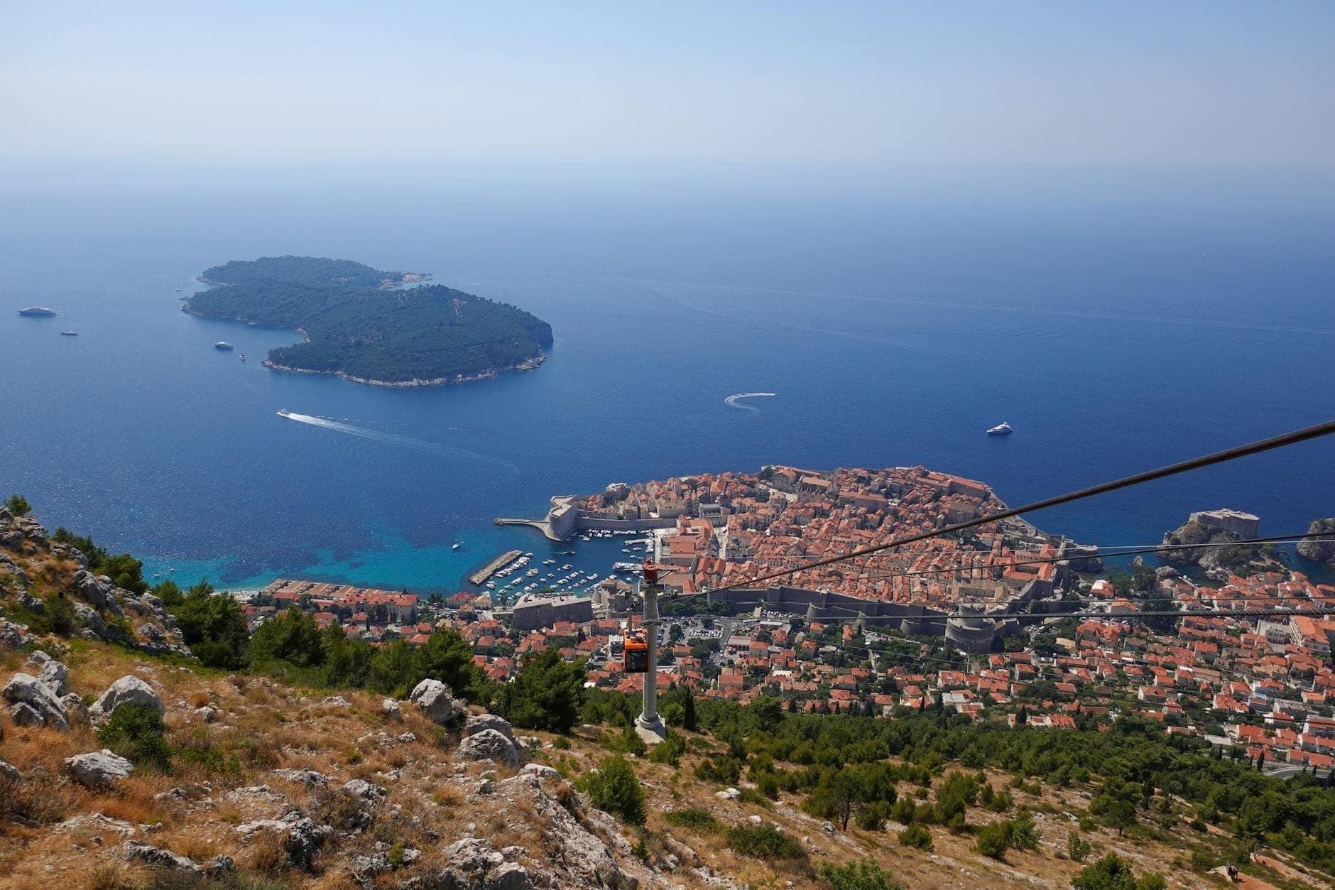 Blick von einem Hügel auf die Altstadt von Dubrovnik, ihre Stadtmauern, die Seilbahn, die Küste, die Insel Lokrum und die Adria an einem klaren Tag.