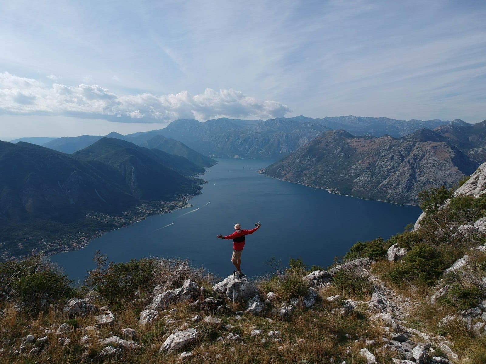 Vue sur la Baie de Kotor, Monténégro, avec une personne en veste rouge debout sur un terrain rocheux surplombant le paysage spectaculaire.