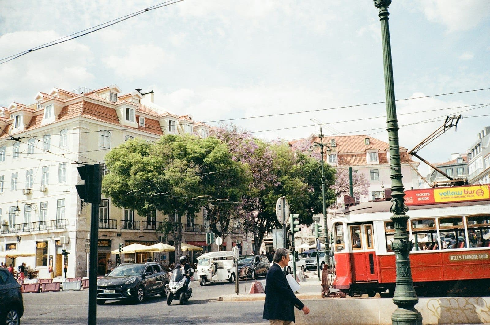 Eléctrico clássico vermelho de Lisboa, carros, motociclos e peões numa movimentada rua do centro com edifícios em tons pastel e árvores ao fundo.