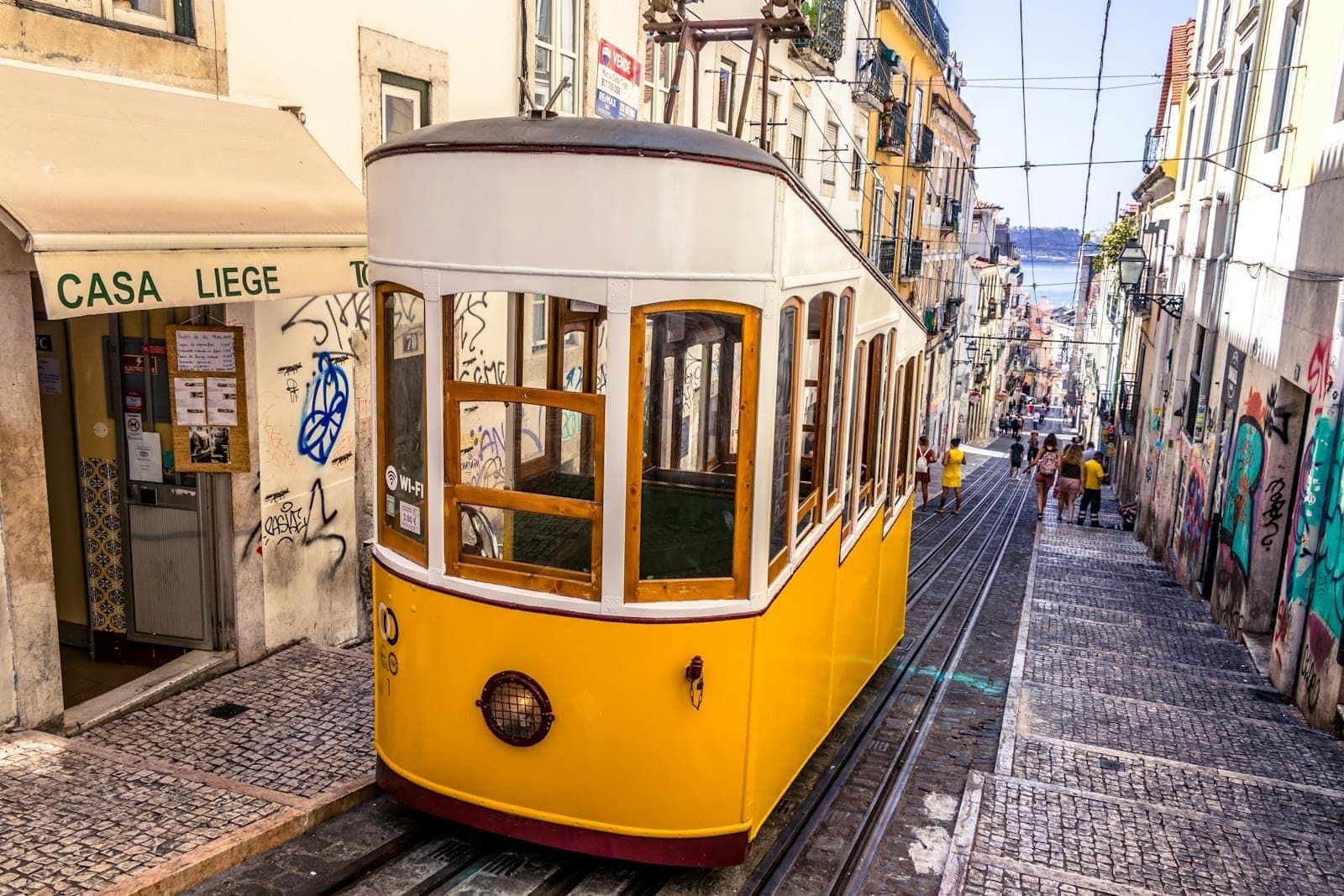 Eléctrico amarelo clássico de Lisboa numa íngreme rua de paralelepípedos ladeada por edifícios coloridos e uma vista do rio ao fundo.