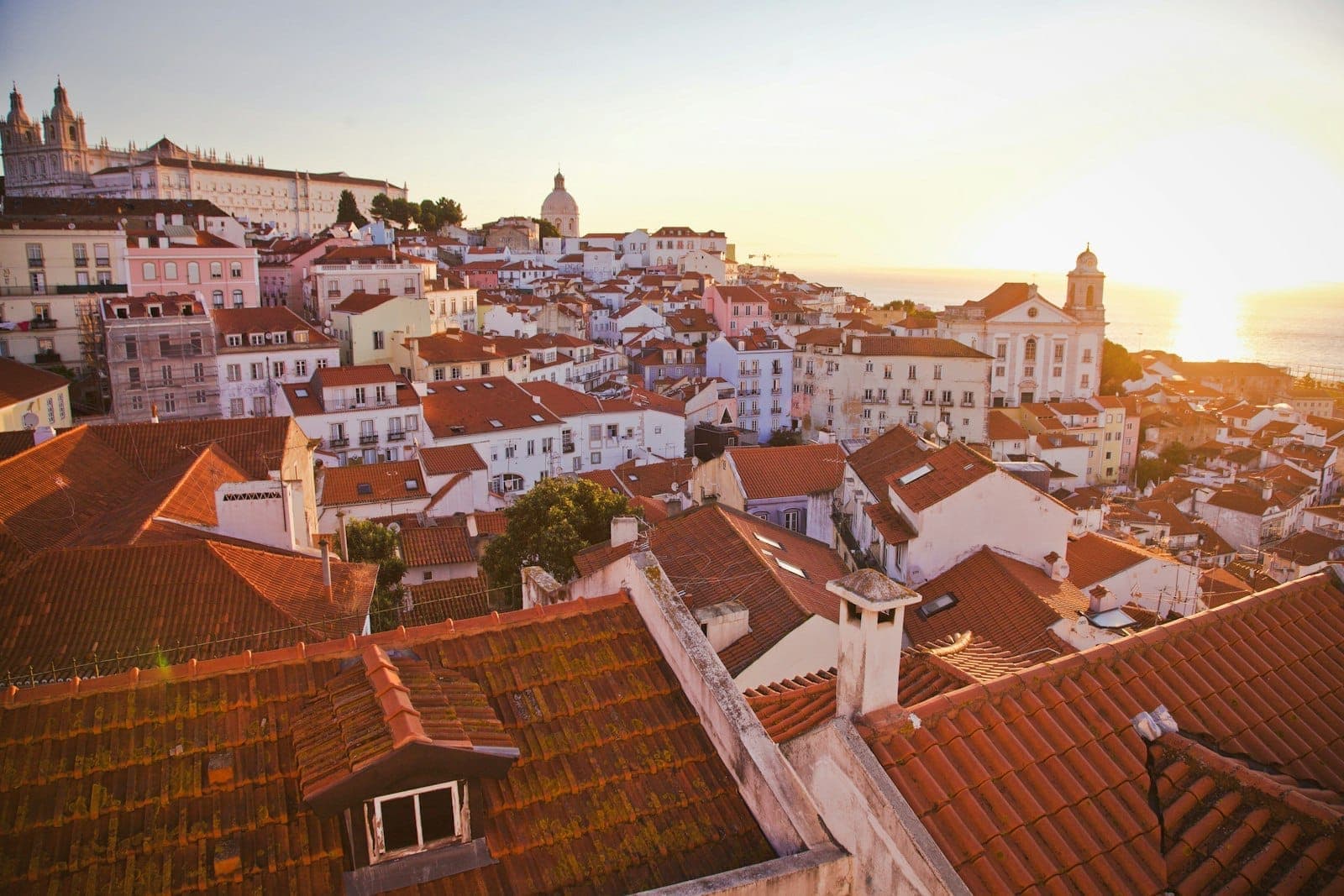 Vista panorâmica sobre os telhados da Alfama em Lisboa e o rio ao amanhecer, com monumentos e cúpula visíveis na luz dourada da hora dourada.