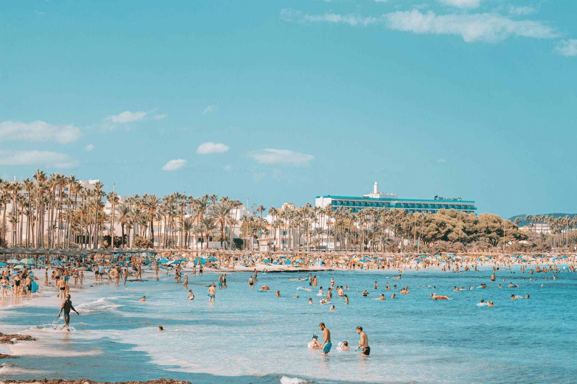 Belebter Strand auf Mallorca mit überfülltem Ufer, vielen schwimmenden und sonnenbadenden Menschen, Palmen und einem großen Resorthotel im Hintergrund.