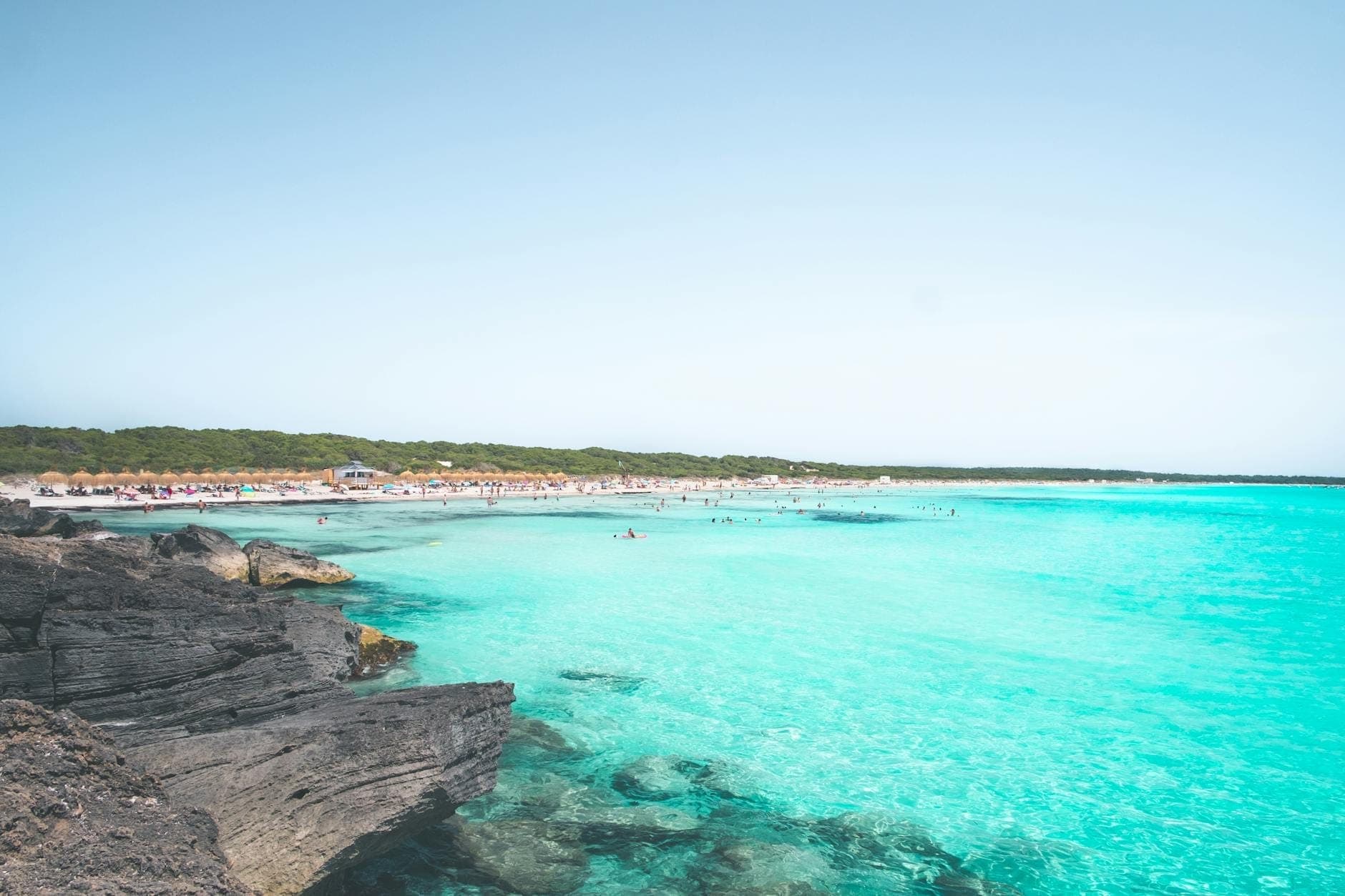 Weitläufiger türkisfarbener Strand mit klarem Wasser, felsigem Vordergrund und Menschen, die sich auf einem langen Sandstreifen in Mallorca sonnen.