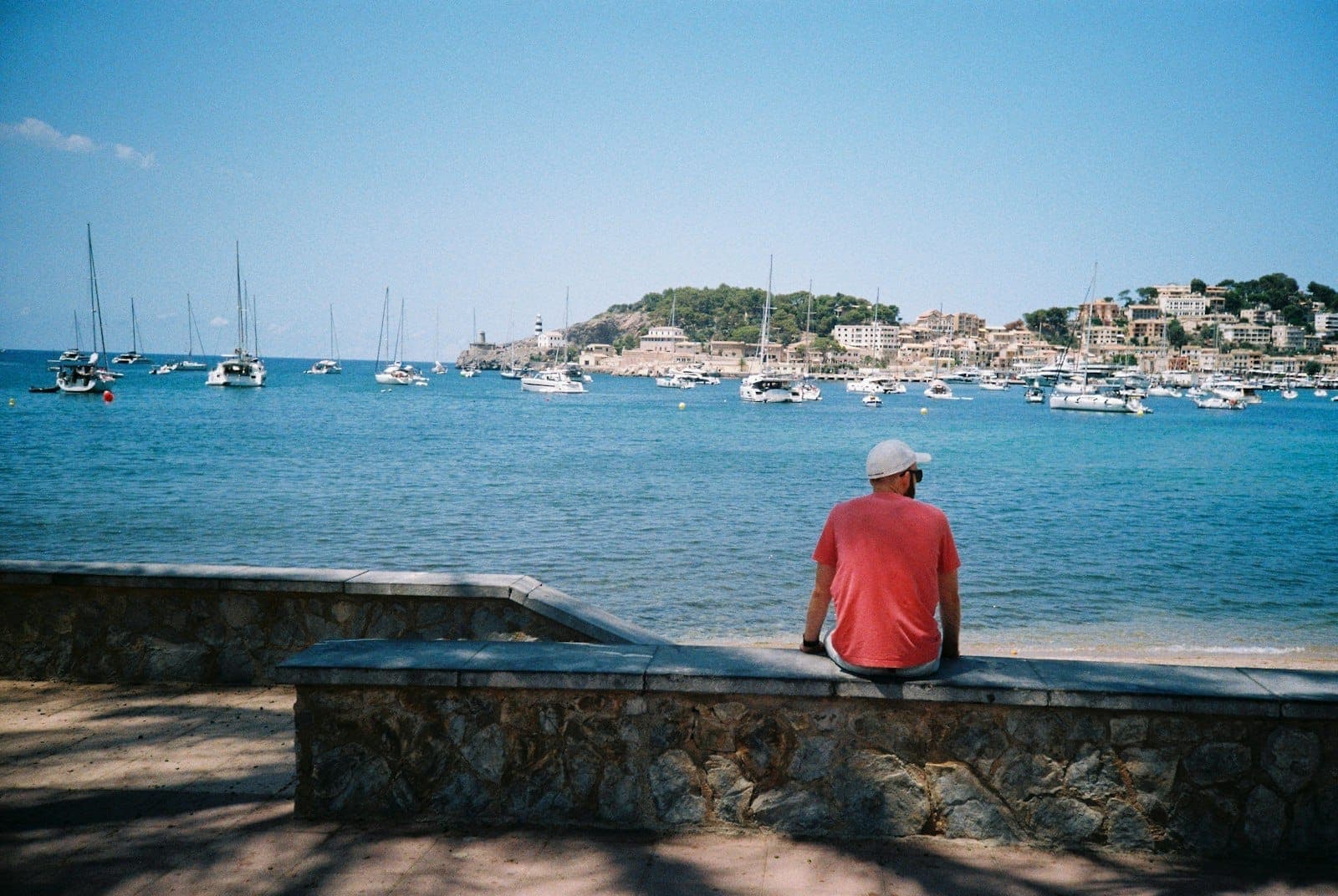 Blick auf die türkisfarbene Bucht von Port de Sóller mit Segelbooten, mediterranen Hanghäusern und einem Leuchtturm unter klarem blauem Himmel.