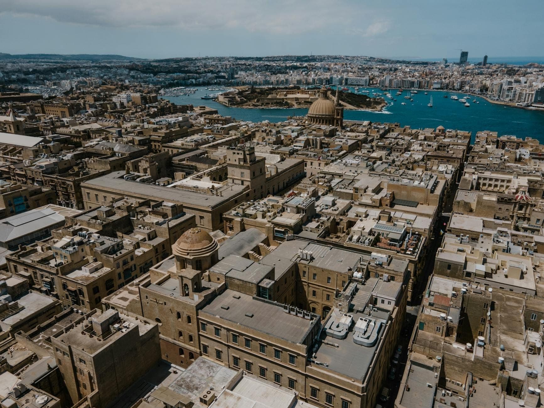 Aerial view of Malta showing historic stone buildings, domed churches, and blue Mediterranean water in the background.