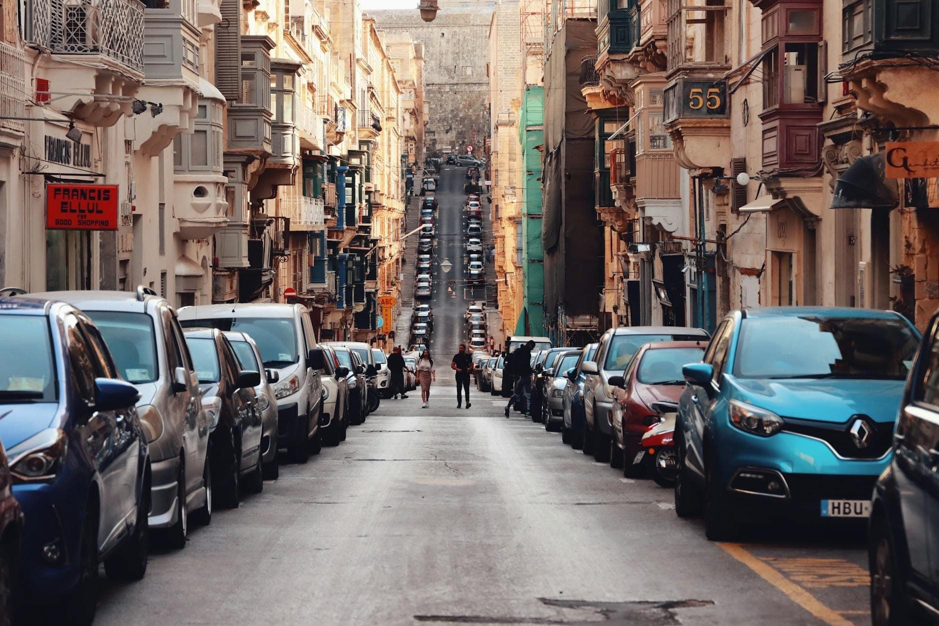A narrow Maltese street crowded with parked cars along both sides and people walking down the center on a hot sunny day.