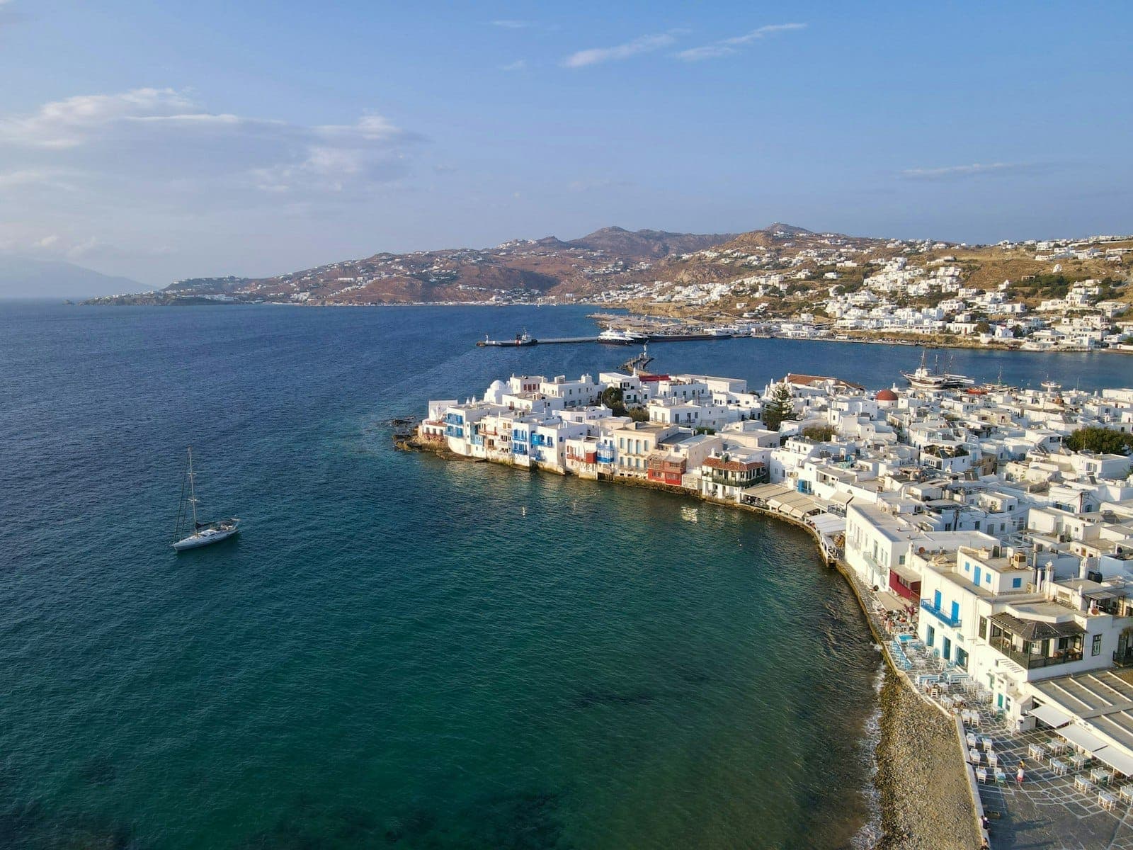 Luftaufnahme der Küste von Mykonos mit weißen Gebäuden, klarem blauem Meer und einem Segelboot, das die ikonische kykladische Architektur und Küstenlandschaft der Insel zeigt.