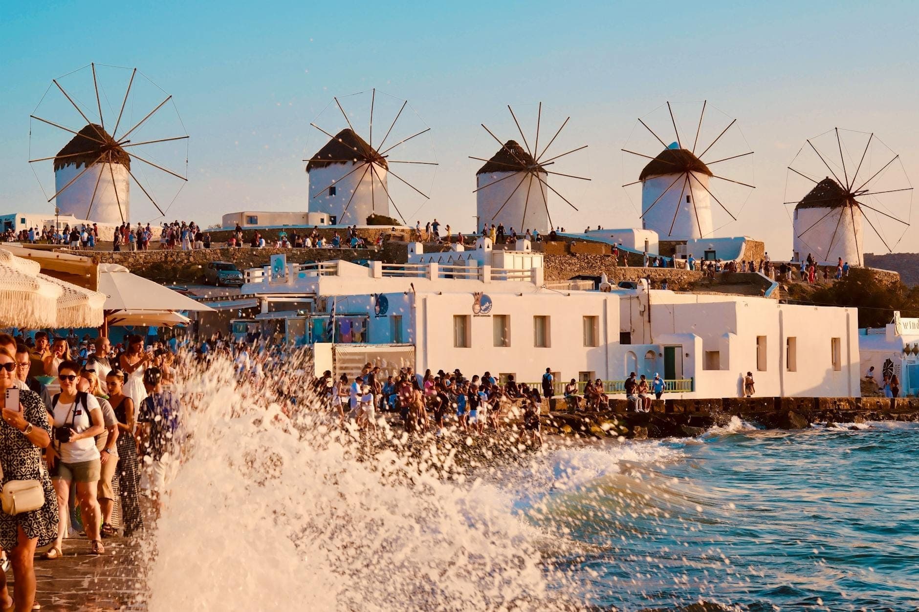 Menschenmassen von Touristen spazieren entlang der Uferpromenade von Mykonos mit den ikonischen Windmühlen im Hintergrund, während Wellen an einem sonnigen Tag gegen das Ufer schlagen.