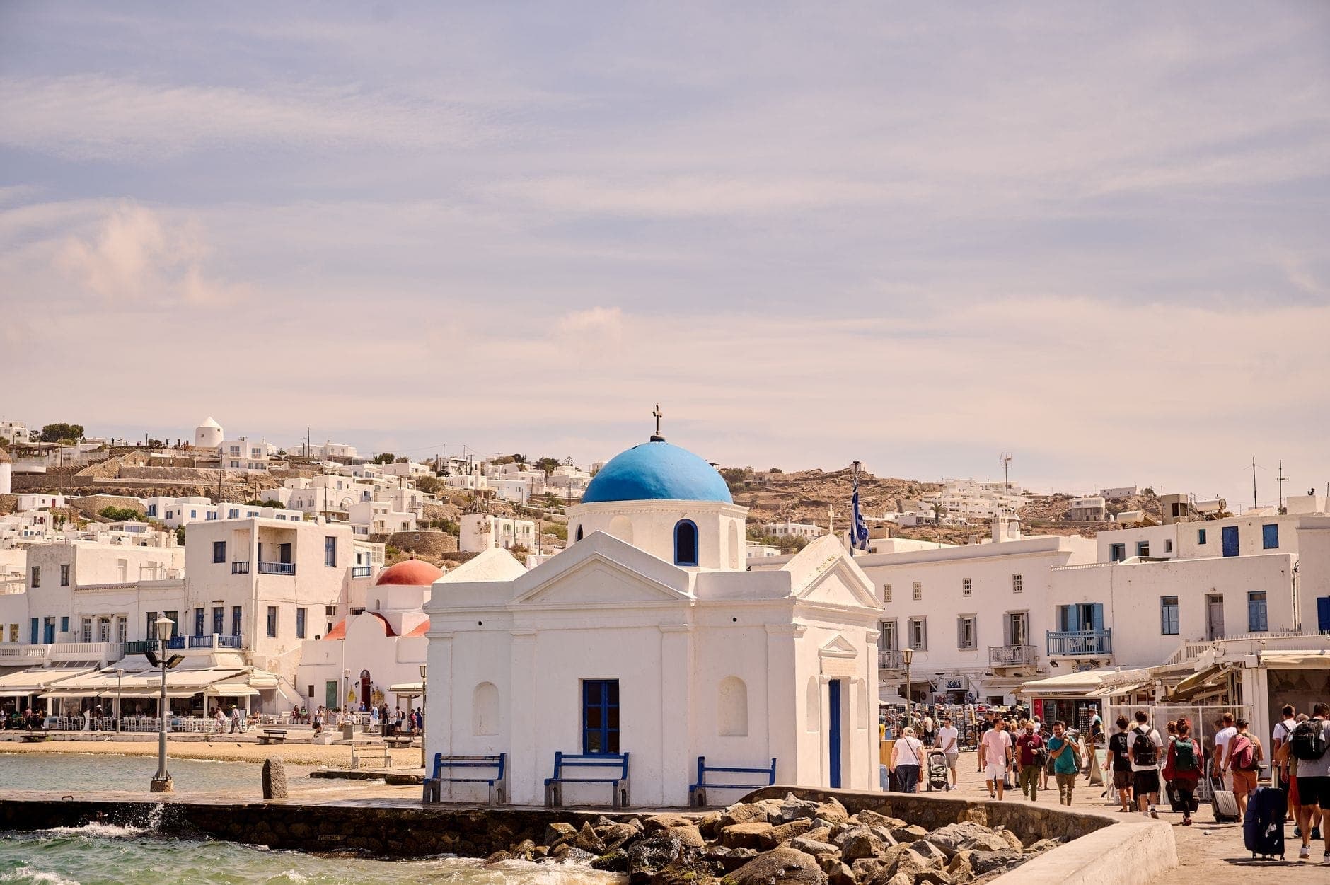 Touristenmassen und Restaurants an der Uferpromenade nahe einer weißen Kirche mit blauer Kuppel in der Stadt Mykonos, die eine belebte und gehobene Sommerszene zeigt.