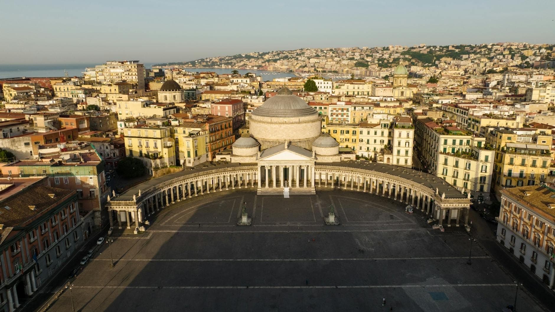 Vista aérea da Piazza del Plebiscito em Nápoles com a Basílica de San Francesco di Paola e coloridos edifícios da cidade ao fundo.
