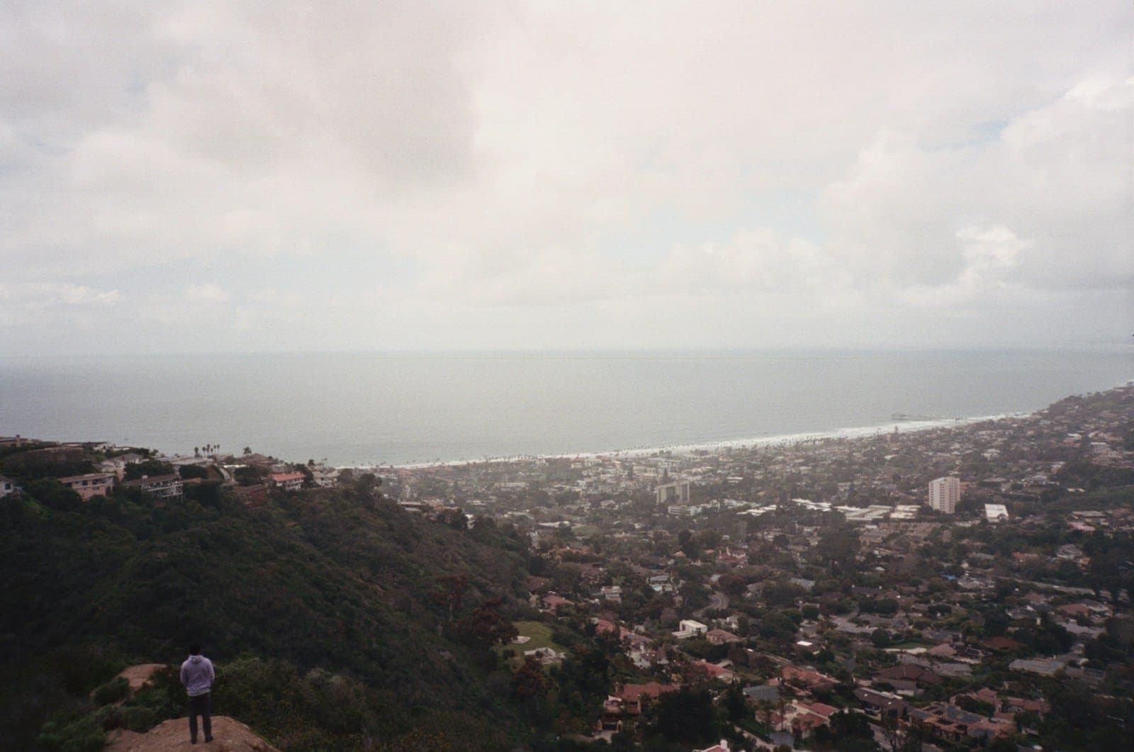 Vue panoramique sur une ville côtière avec collines, maisons éparses et mer à l'horizon sous un ciel nuageux.