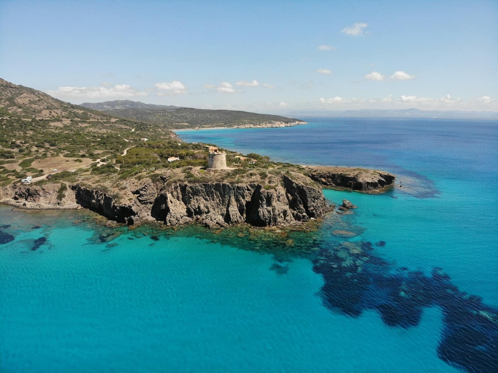 Vista aérea de la costa de Cerdeña con aguas turquesa, acantilados escarpados y una histórica torre de piedra en el promontorio.