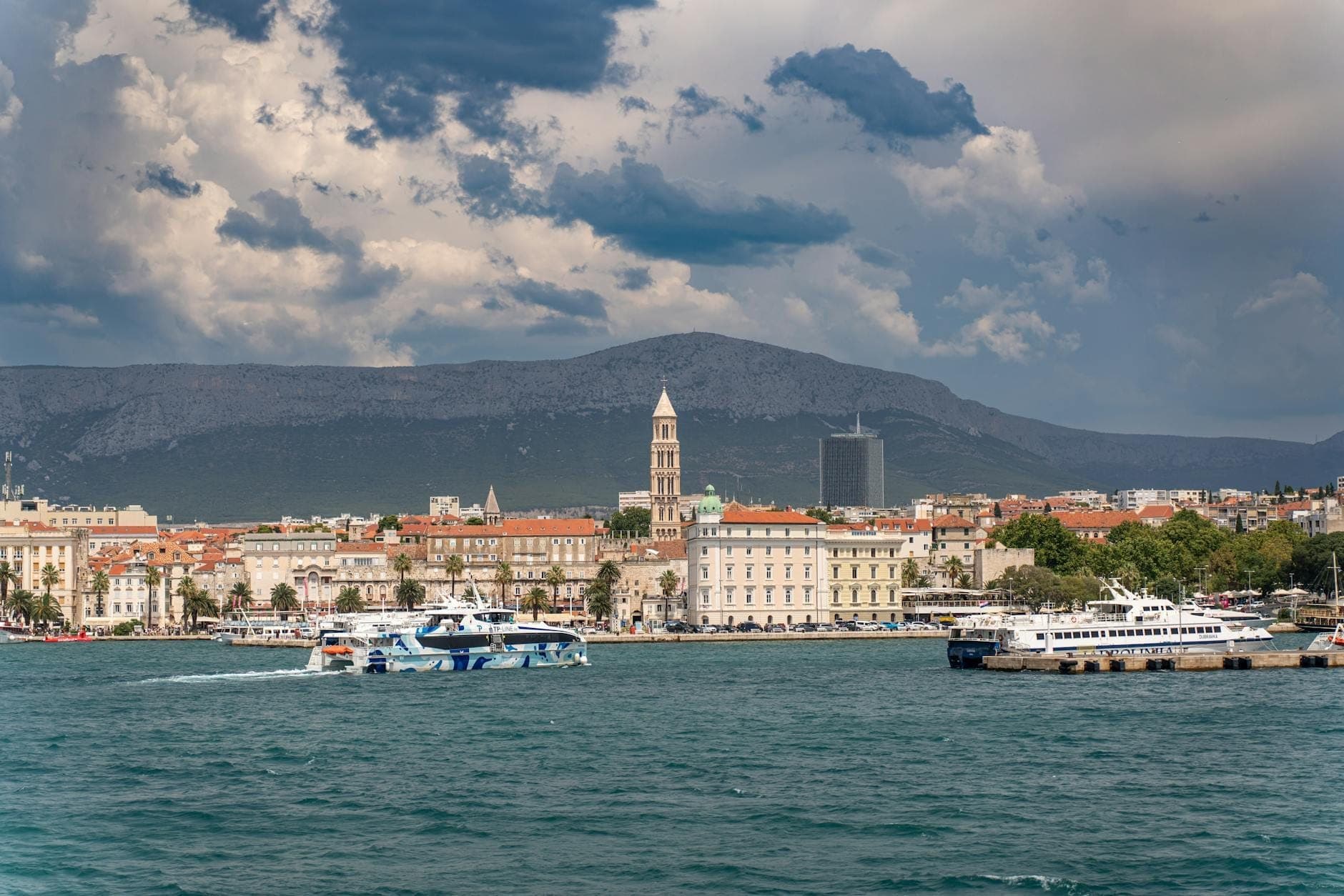 Vista del lungomare di Spalato con barche, edifici della città e il campanile della cattedrale sotto un cielo minaccioso, che evidenzia un'atmosfera costiera urbana.