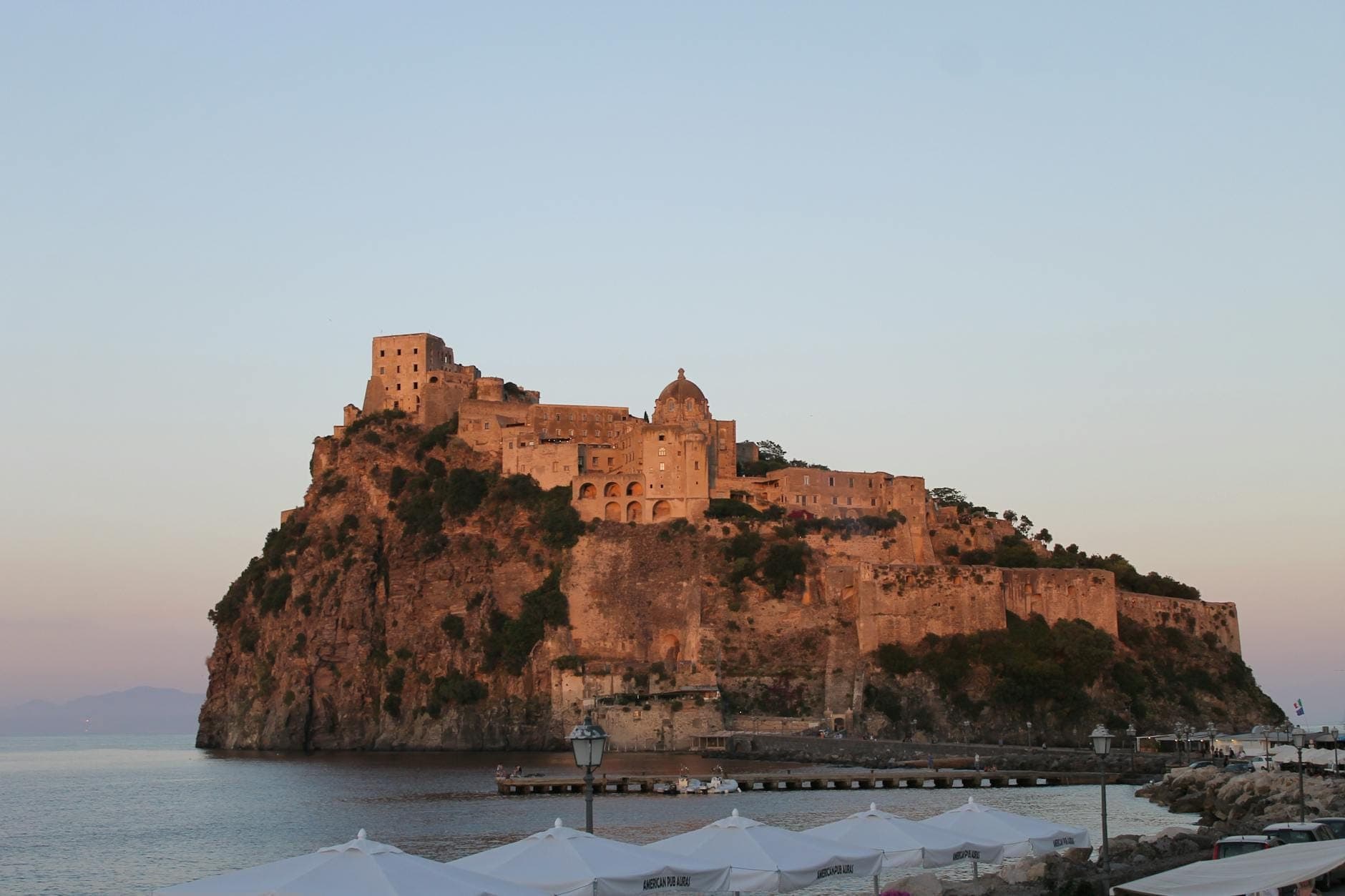 O Castelo Aragonês sobre um ilhéu rochoso na costa de Ischia ao pôr do sol, com águas calmas e um céu em tons pastéis ao fundo.