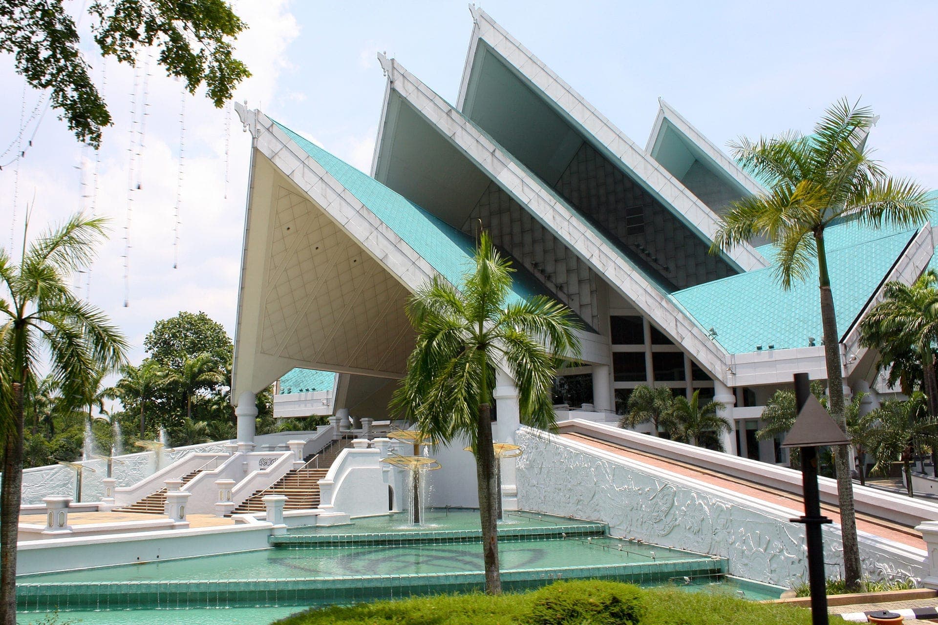 Arquitetura moderna impressionante do Istana Budaya com telhados angulares turquesa, escadarias brancas, palmeiras e espelhos d'água sob luz do dia, Kuala Lumpur.