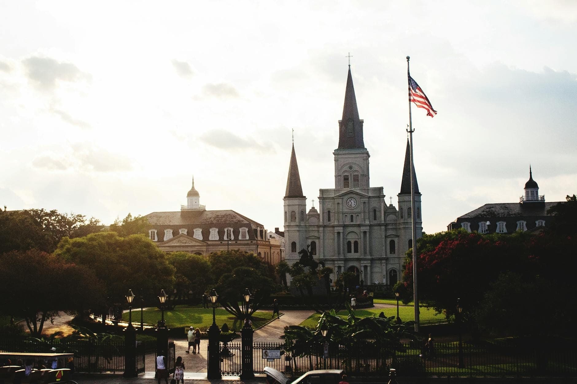 A wide view of Jackson Square showing the St. Louis Cathedral at center, surrounded by lush gardens, historic buildings, and an American flag at sunset.
