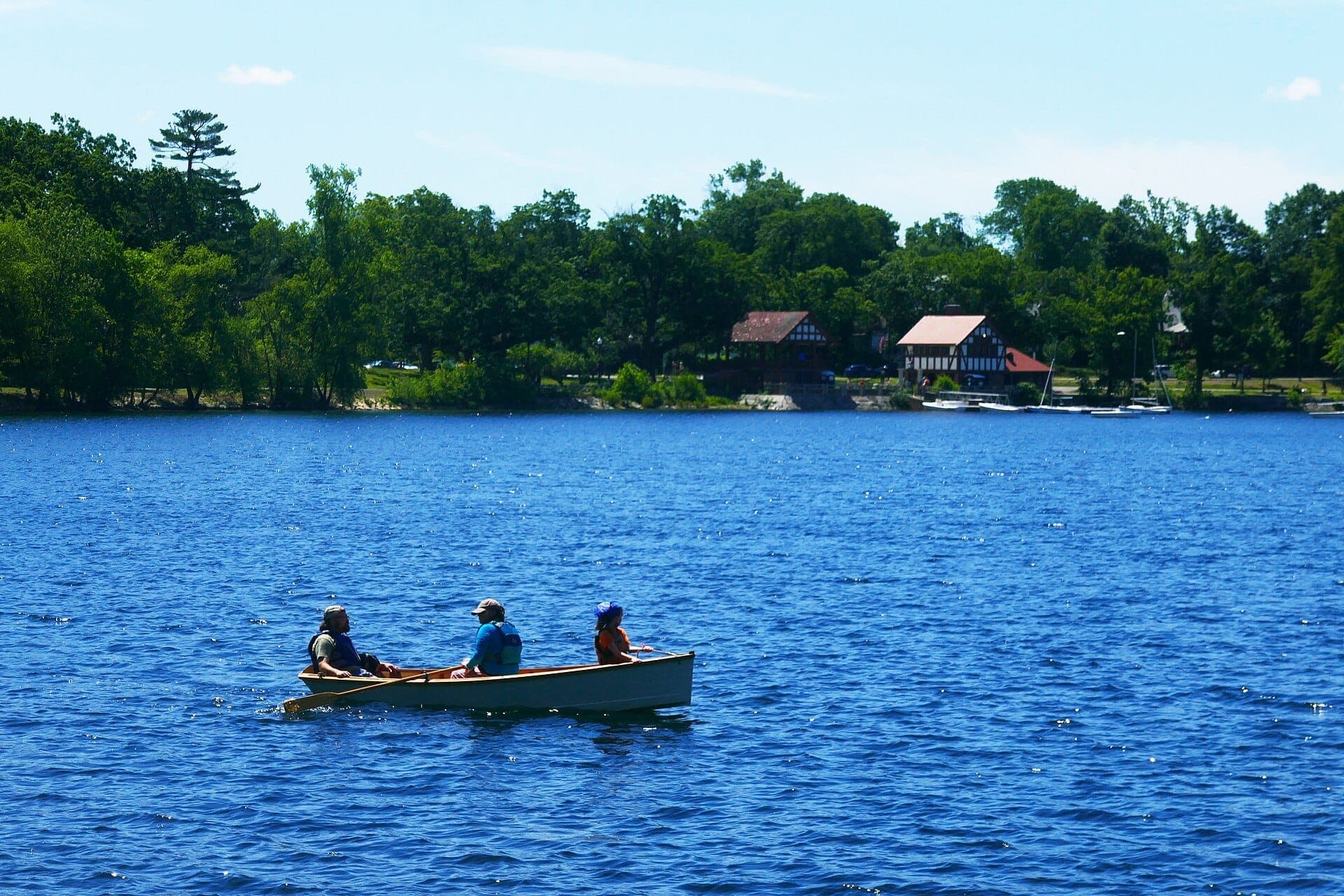 Trois personnes rament dans une petite barque sur Jamaica Pond avec des arbres luxuriants et les bâtiments du hangar à bateaux en arrière-plan par une journée claire.