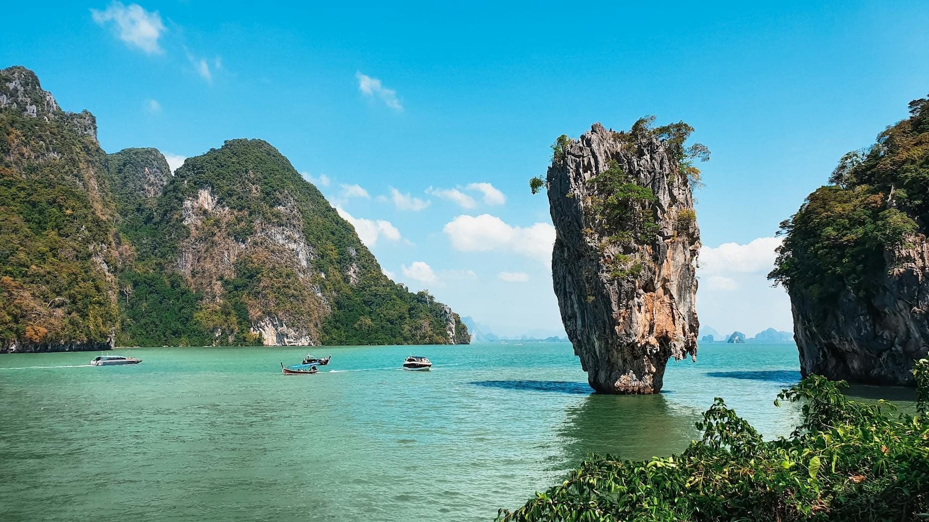 Iconic limestone rock of James Bond Island rising from emerald water, surrounded by green cliffs and boats under a bright blue sky in Phang Nga Bay, Thailand.