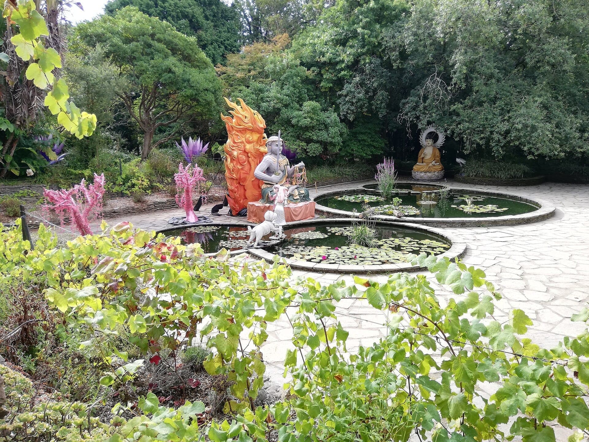 Kreisförmige Seerosenteiche mit bunten Statuen, darunter ein Buddha, umgeben von üppigem Grün im Jardim Botânico do Porto an einem hellen Tag.