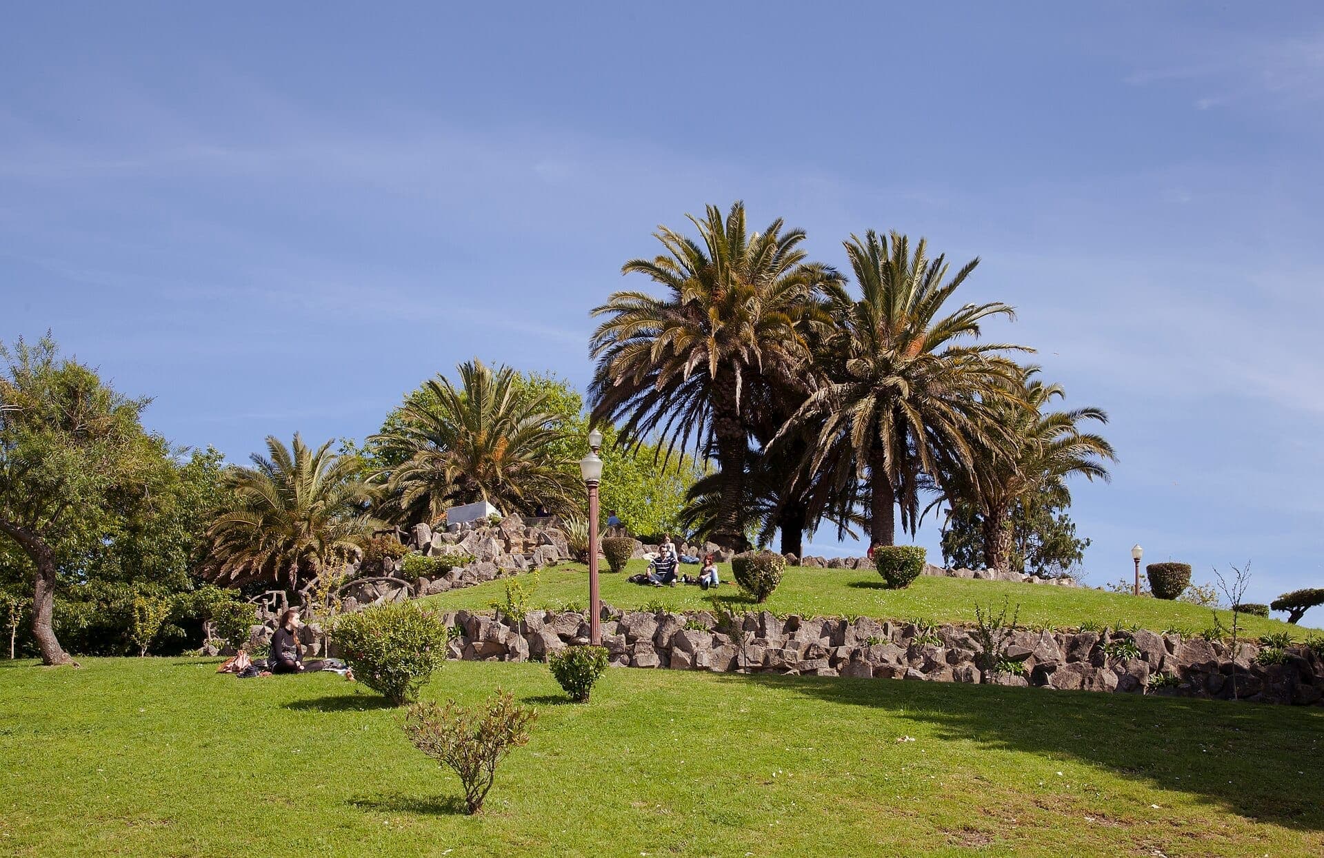 Menschen entspannen sich auf den grünen Rasenflächen des Jardim do Morro, umgeben von Palmen und Steinterrassen unter einem klaren blauen Himmel.