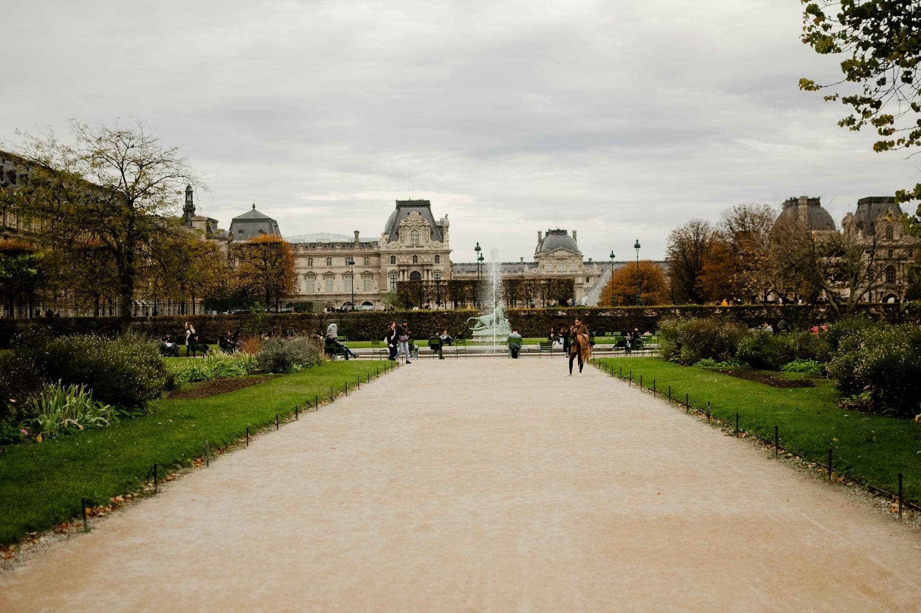 Wide pathway in the Jardin des Tuileries with green lawns, flower beds, people strolling, a fountain at the center, and the Louvre in the background under a cloudy sky.