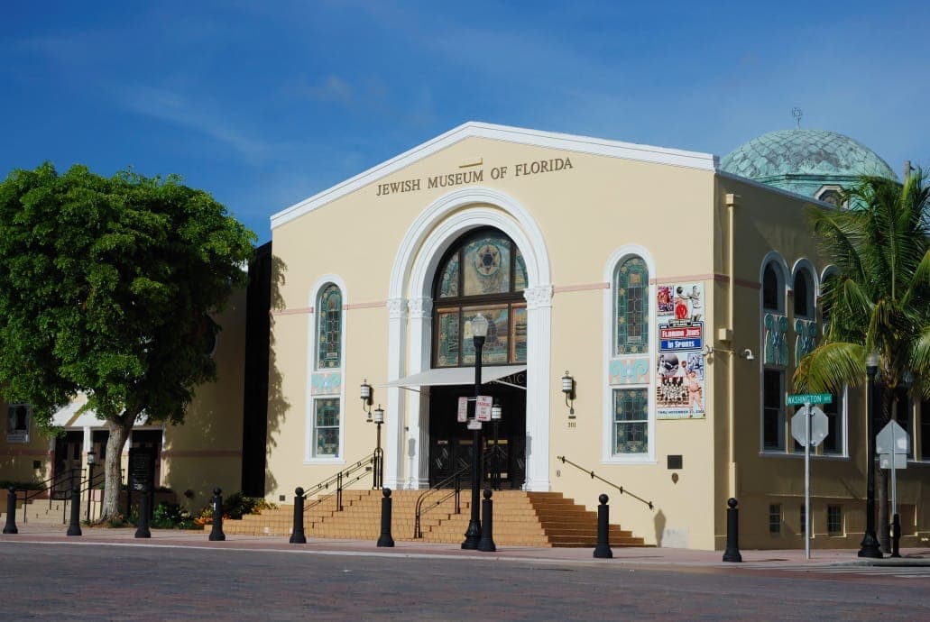 Front view of the Jewish Museum of Florida–FIU, showing its yellow facade, stained-glass windows, and prominent copper dome under a clear blue sky.