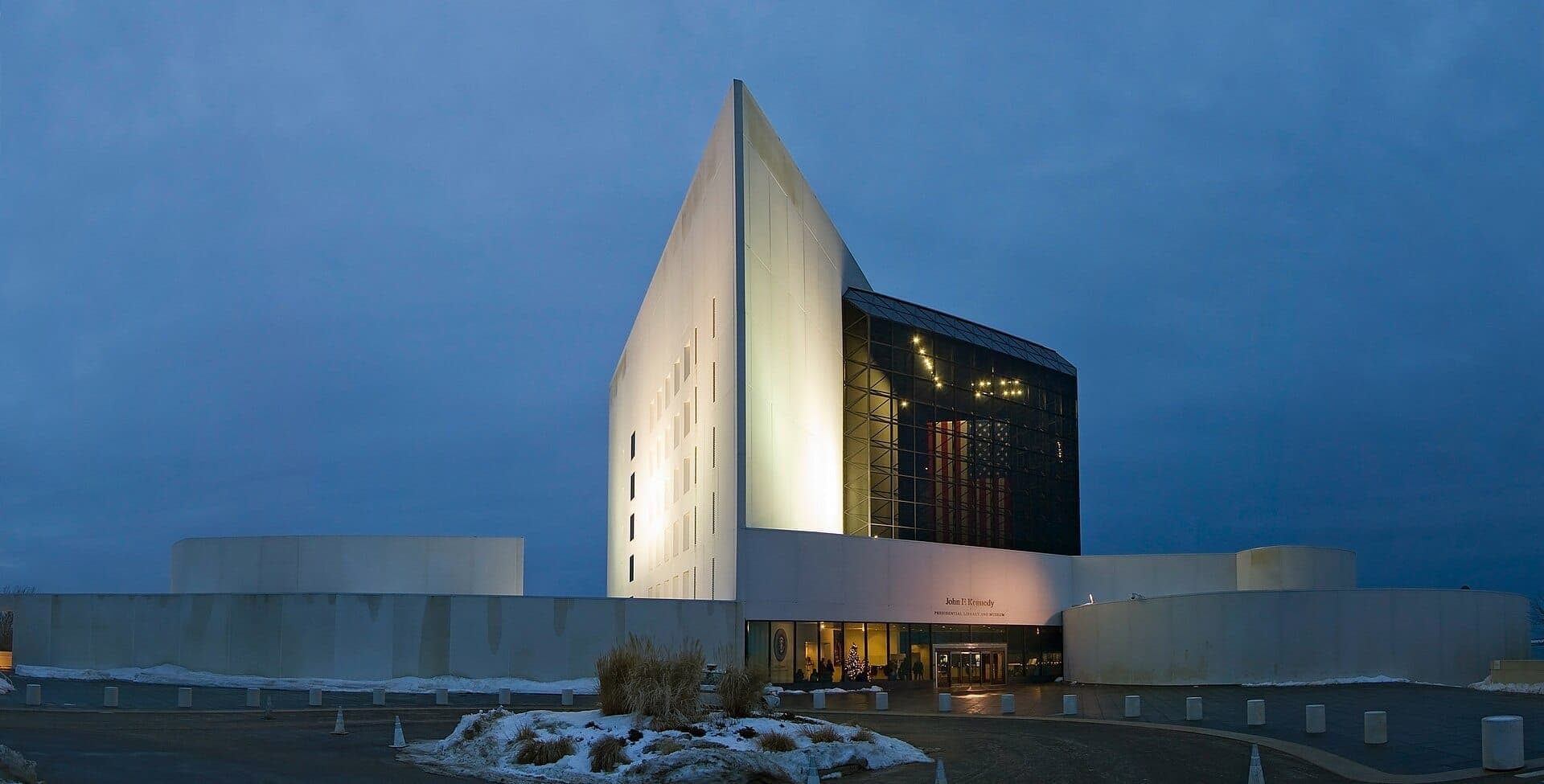 La Bibliothèque et Musée Présidentiel John F. Kennedy au crépuscule, avec une architecture moderne illuminée, une façade en verre et des plaques de neige sur le sol.