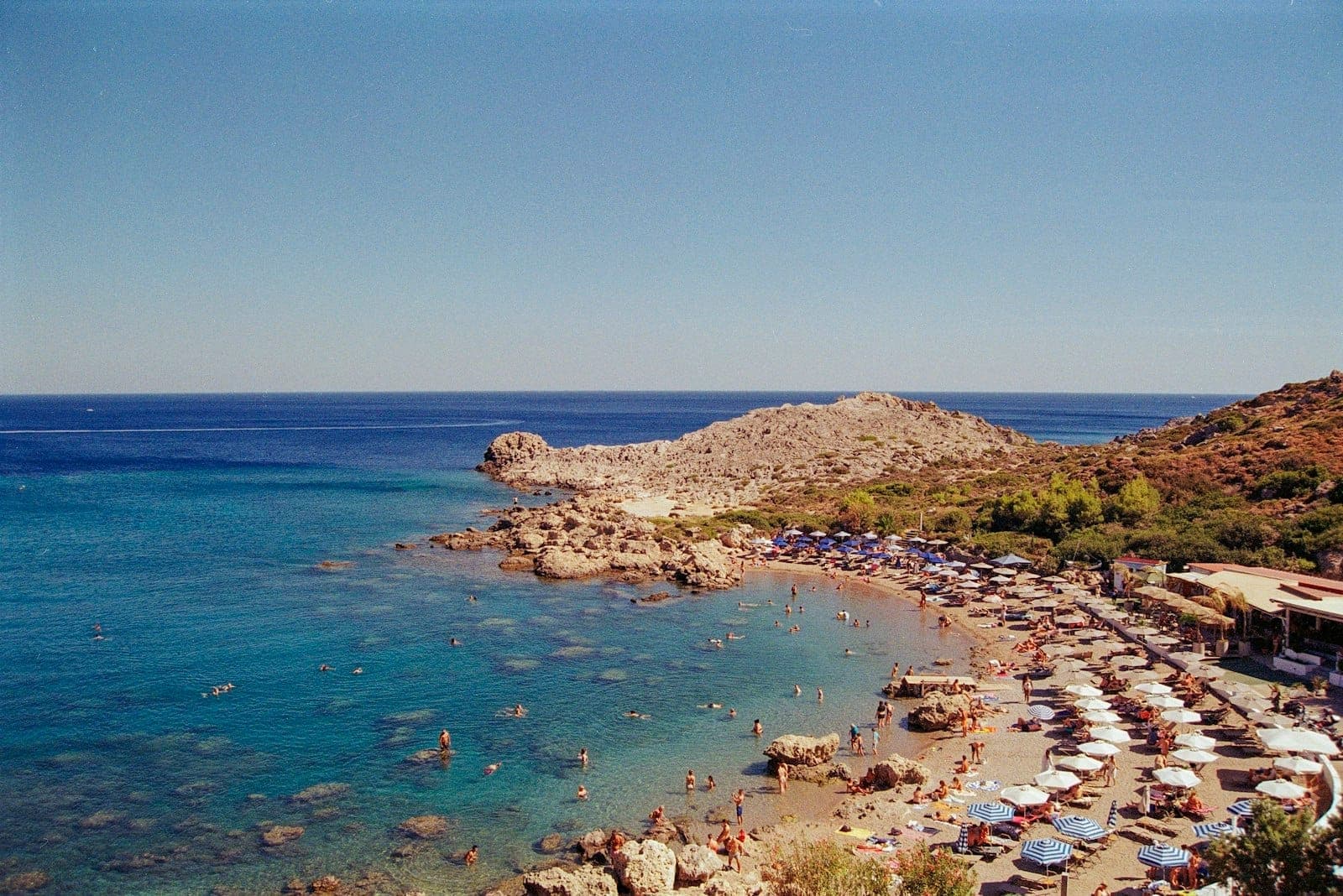 Plage animée de Kallithea avec rangées de parasols et de baigneurs, côte rocheuse, mer d'un bleu cristallin et douces collines sous un ciel dégagé.