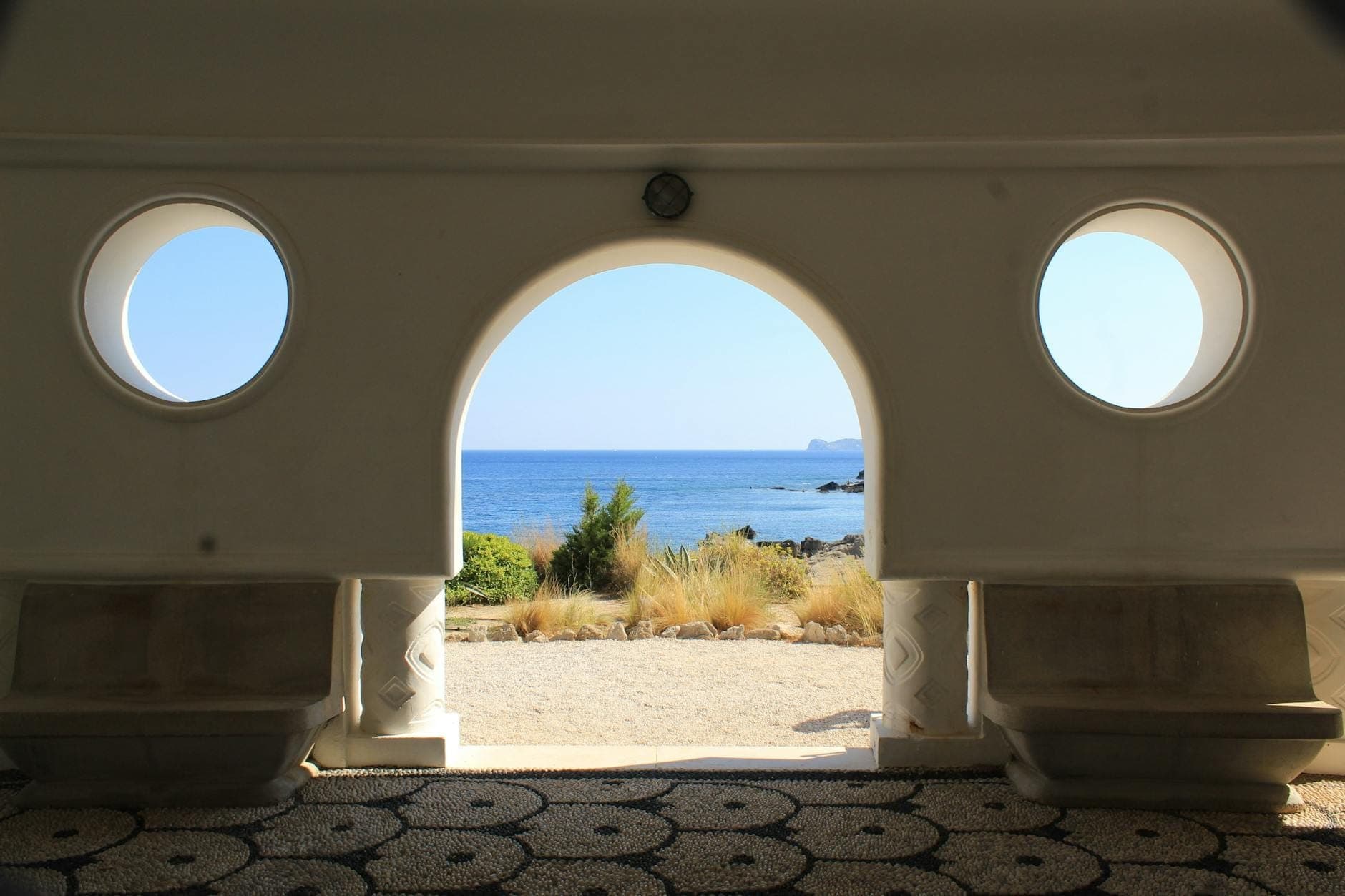 Vue à travers une porte Art Déco cintrée aux fenêtres rondes, donnant sur la mer bleue et le rivage de Kallithea Springs à Rhodes.