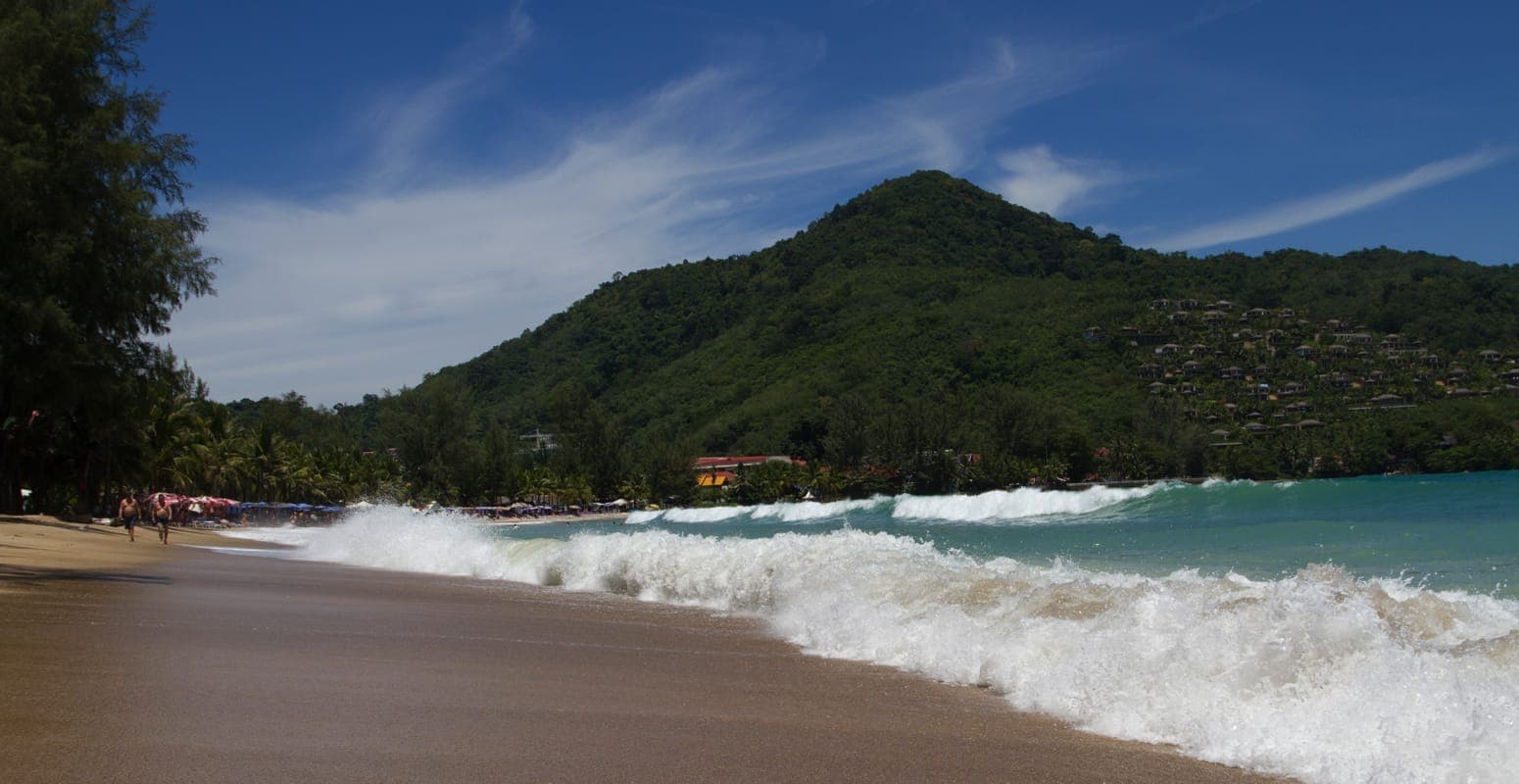 Waves crash onto the golden sands of Kamala Beach in Phuket, with lush green hills and seaside buildings in the background under a blue sky.