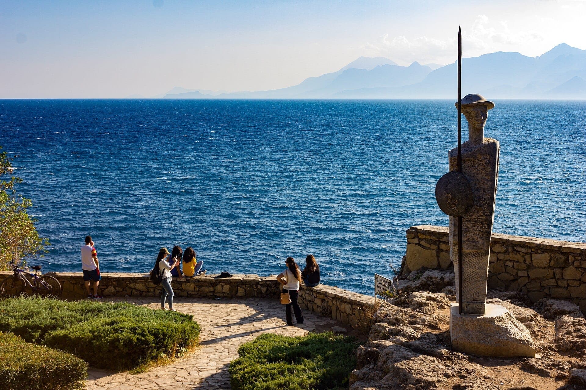 Malerischer Blick vom Karaalioğlu-Park mit einem Steinweg, einer Statue am Meer und Besuchern mit Blick auf das strahlend blaue Mittelmeer.