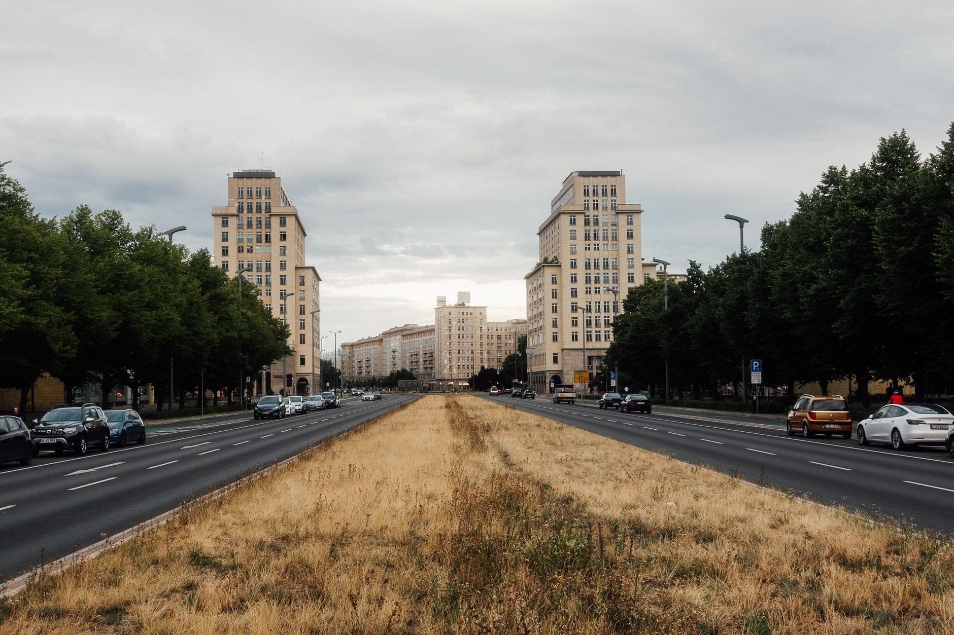 ภาพมุมกว้างของ Karl-Marx-Allee ในเบอร์ลิน กับตึกสถาปัตยกรรมสตาลิน สองข้างทางเป็นอาคารสูงรถยนต์จอดเรียงและไม้ใหญ่ใต้ท้องฟ้าครึ้ม