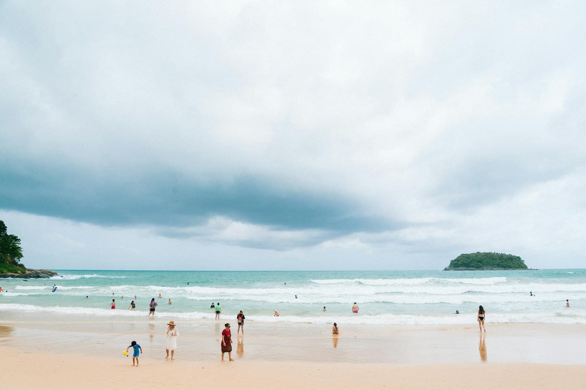People enjoying the surf and sand at Kata Yai Beach, Phuket, with turquoise water, white sand, and a small island under a dramatic sky.