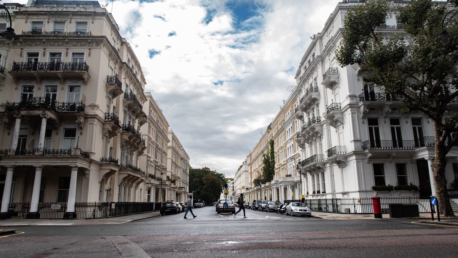 Vista de una amplia calle con elegantes edificios blancos de fachadas escalonadas, clásicas rejas negras y algunos peatones cruzando bajo un cielo dramáticamente nublado en Kensington & Chelsea.