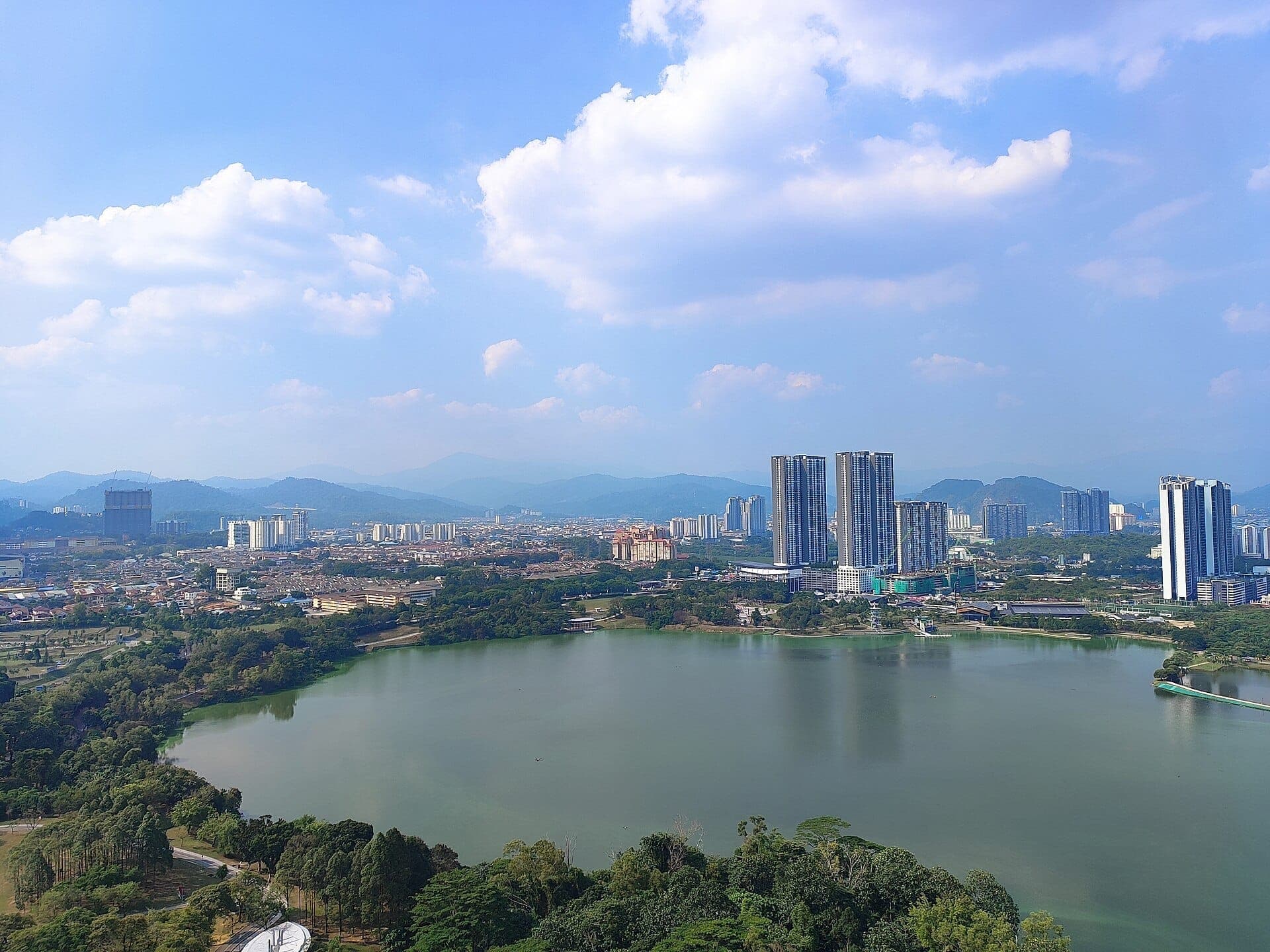 Kepong Metropolitan Park'ın büyük gölünün çevre ağaçları, şehir gökdelenleri ve dağlarıyla parlak mavi gökyüzü altındaki gündüz panoramik görünümü.