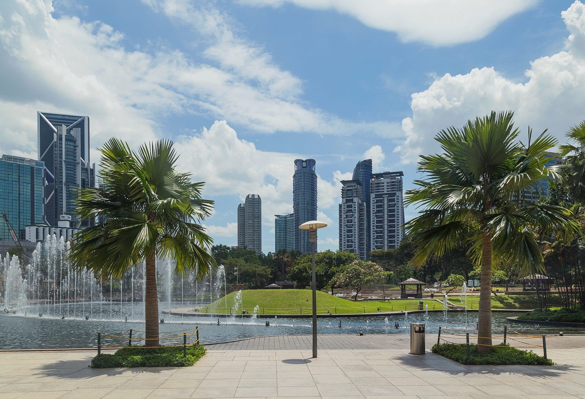 KLCC Park'ın palmiye ağaçları, bir lagün çeşmesi, yeşil çimen ve parlak mavi gökyüzü altında Kuala Lumpur gökdelenleriyle geniş görünümü.