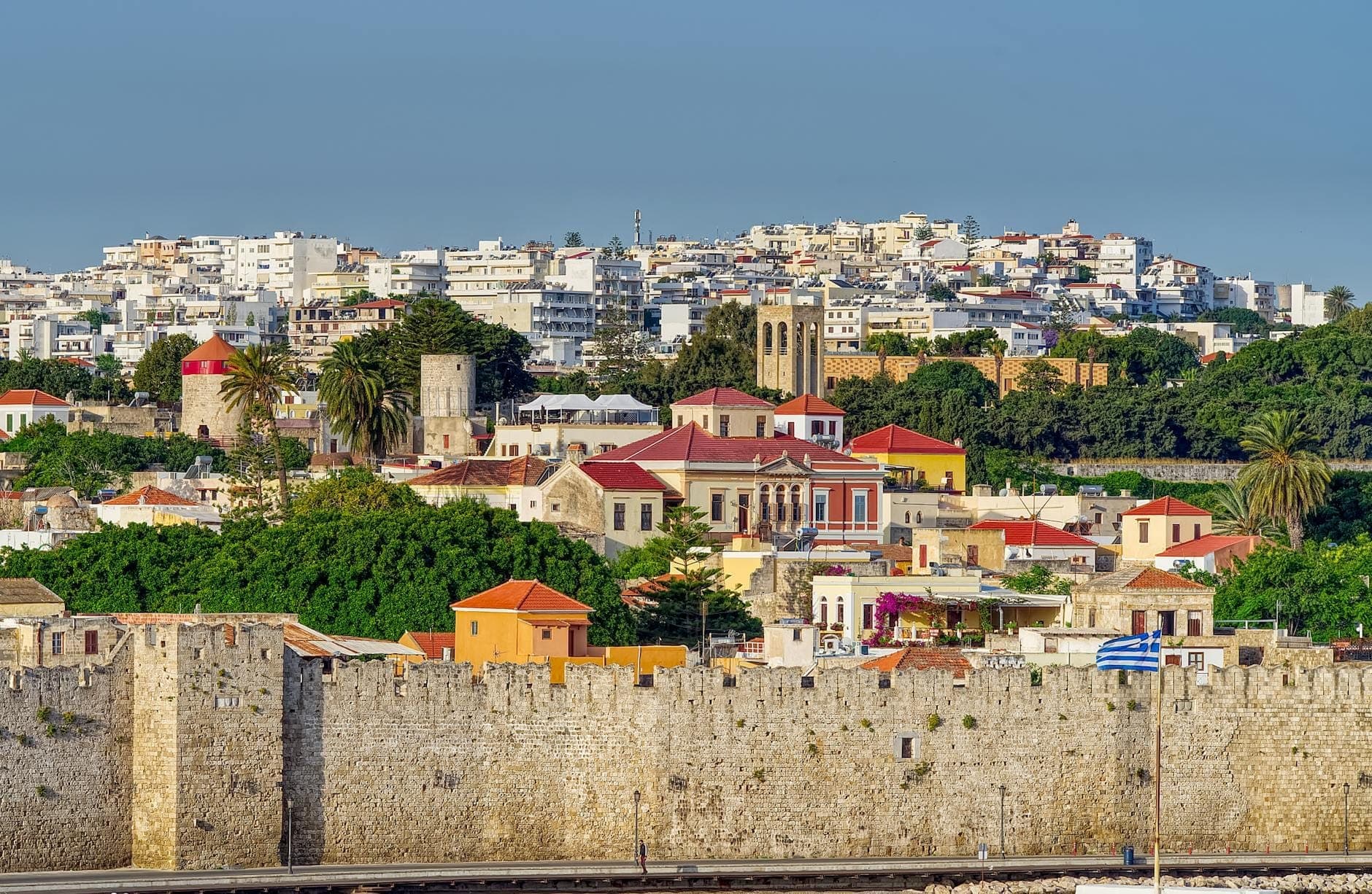 View of the medieval stone walls and historic buildings of Rhodes Old Town with modern city in the background under clear sky.