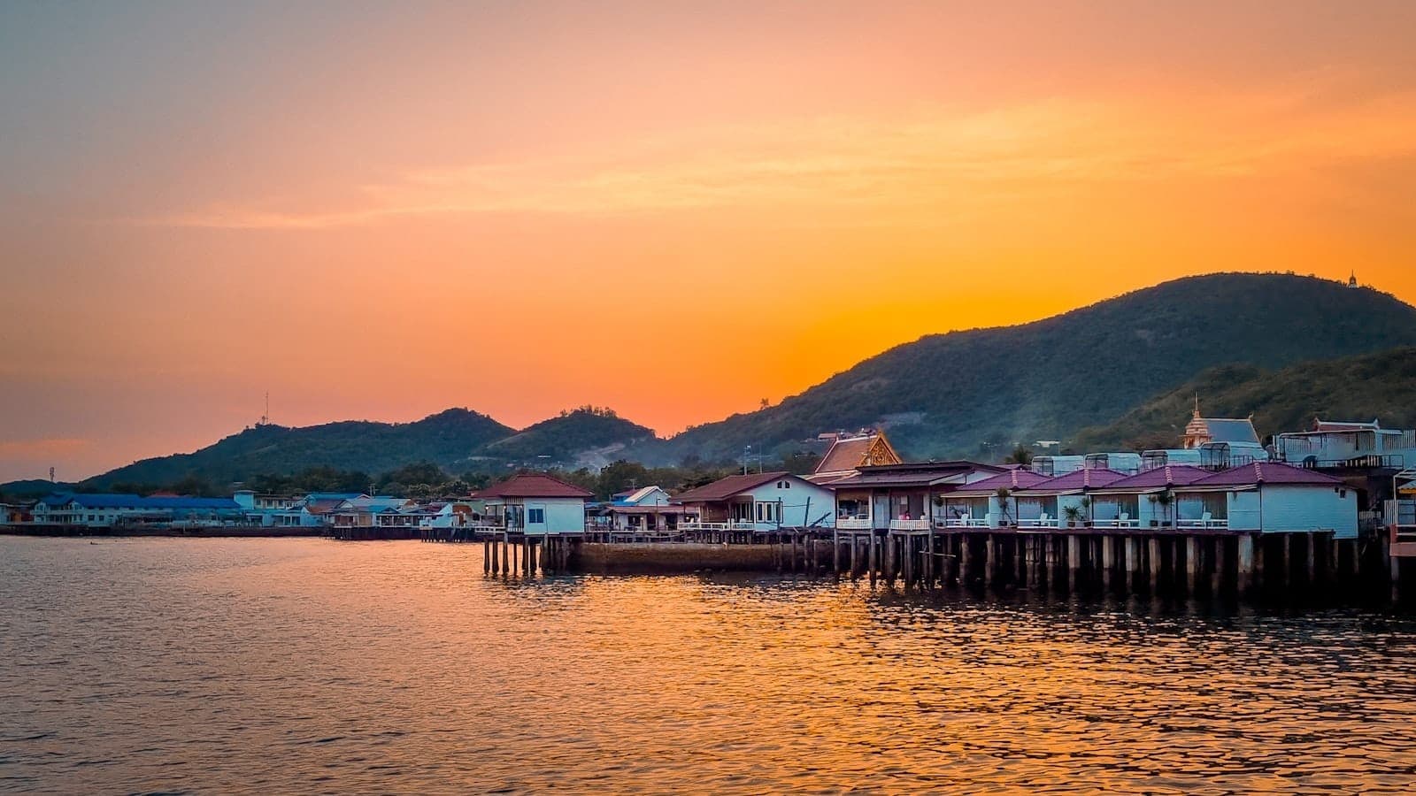 Colorful waterfront houses on stilts at Koh Larn during a vibrant orange sunset, with green hills rising in the background.