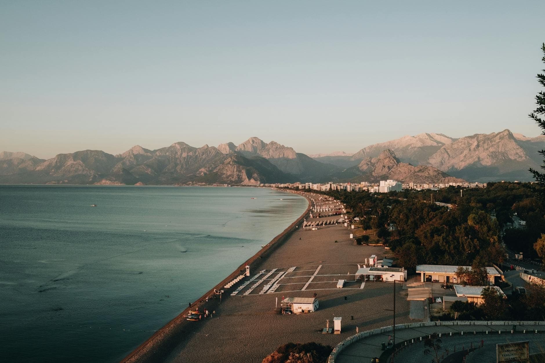 Ein langer Abschnitt des Konyaaltı Strands mit ruhigem blauem Meer, Kiesufer, Gebäuden und den beeindruckenden Taurusbergen im Hintergrund unter einem klaren Abendhimmel.