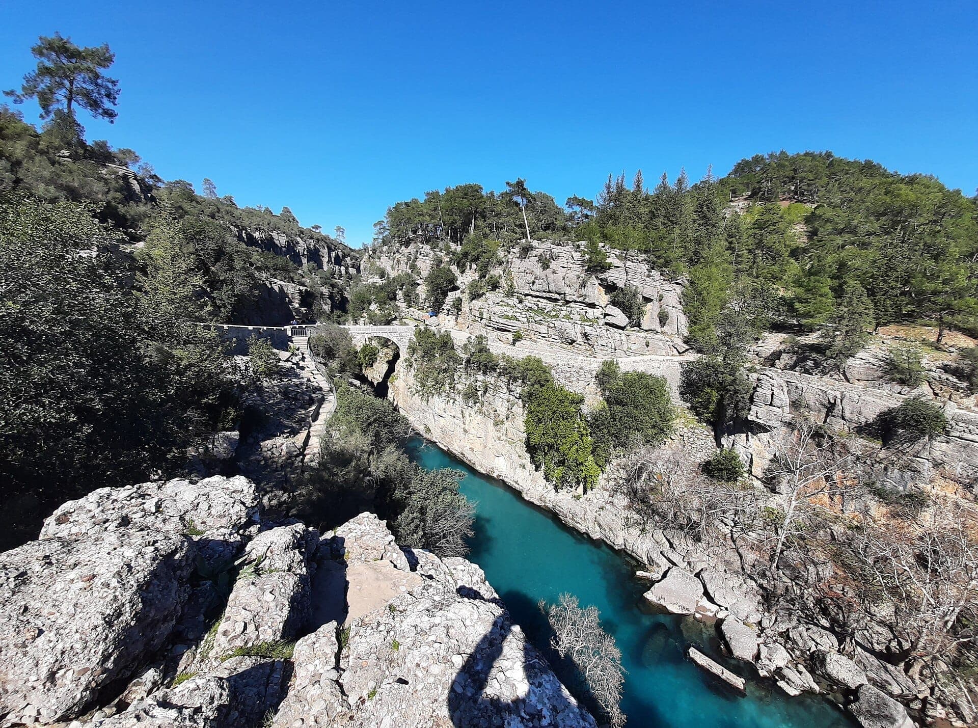 Eine alte Steinbrücke überspannt den türkisfarbenen Köprülü-Fluss in einer felsigen Schlucht, umgeben von dichtem grünen Wald unter einem klaren blauen Himmel.