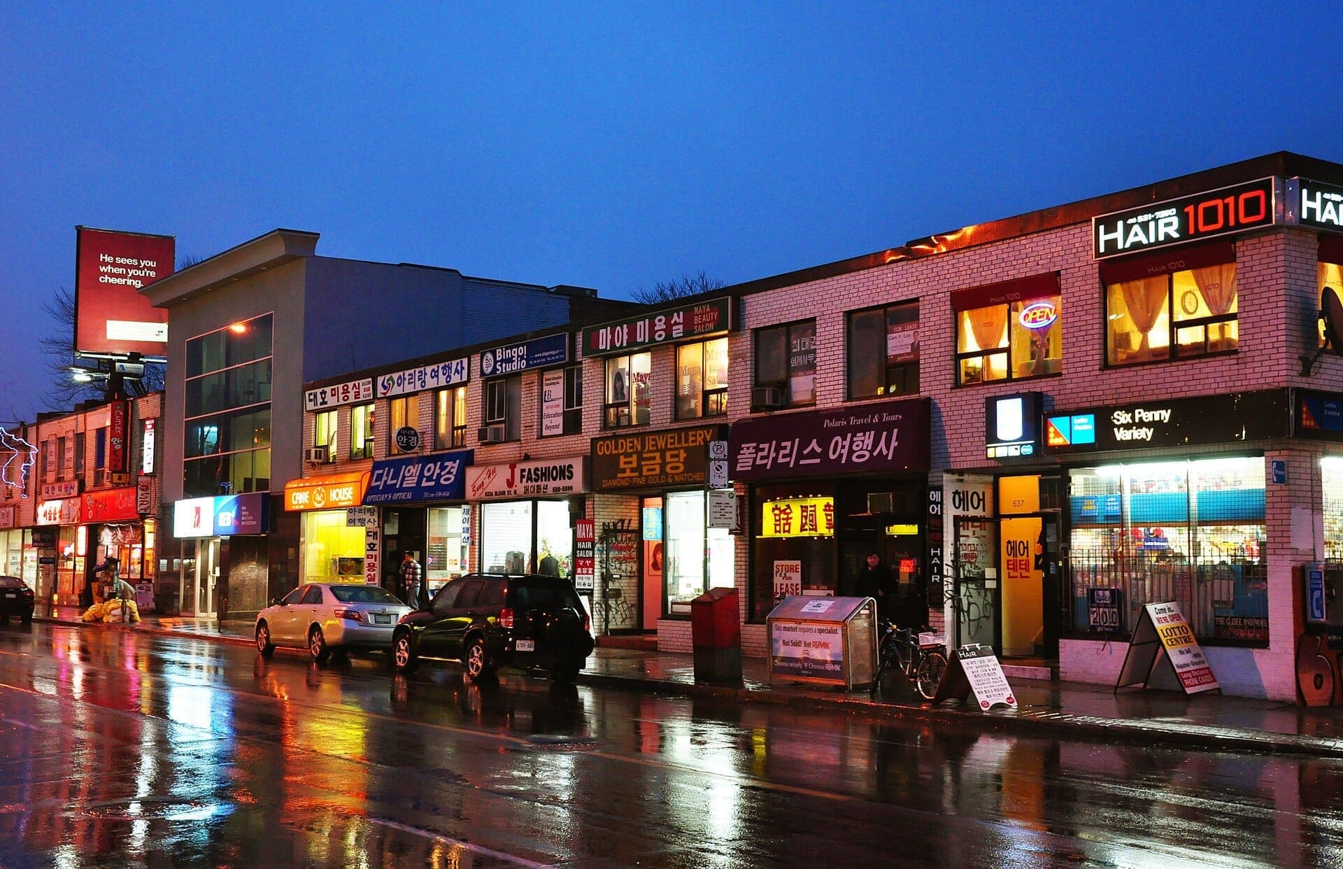 Negozi con insegne in coreano e luci al neon fiancheggiano una Bloor Street bagnata e riflettente nel Koreatown di Toronto al crepuscolo.